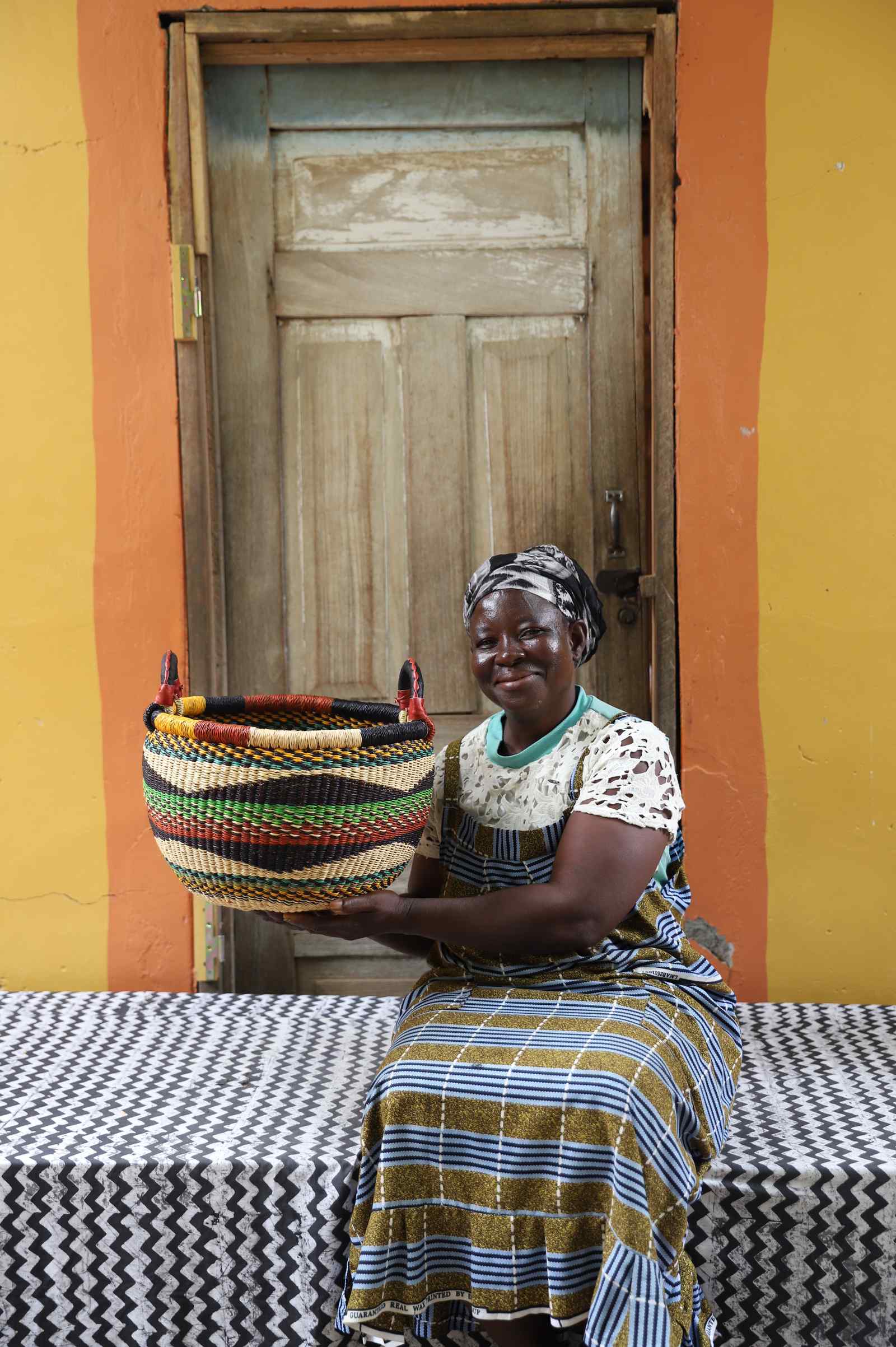Small handwoven Bolga Gbenka storage basket in multicolour bands, styled beside a seated woman in olive.