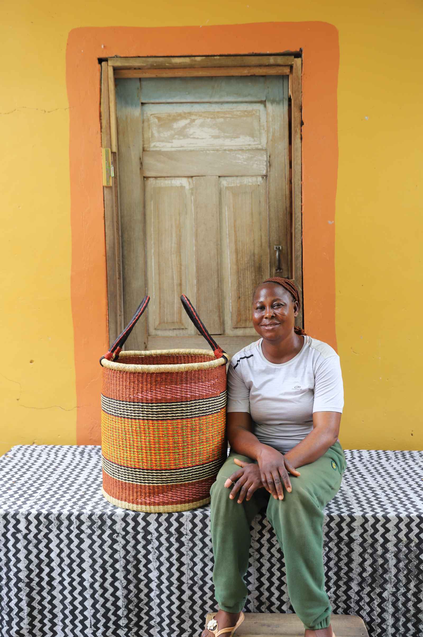 Medium handwoven Bolga laundry basket in orange, yellow and red bands, beside a seated woman in olive.