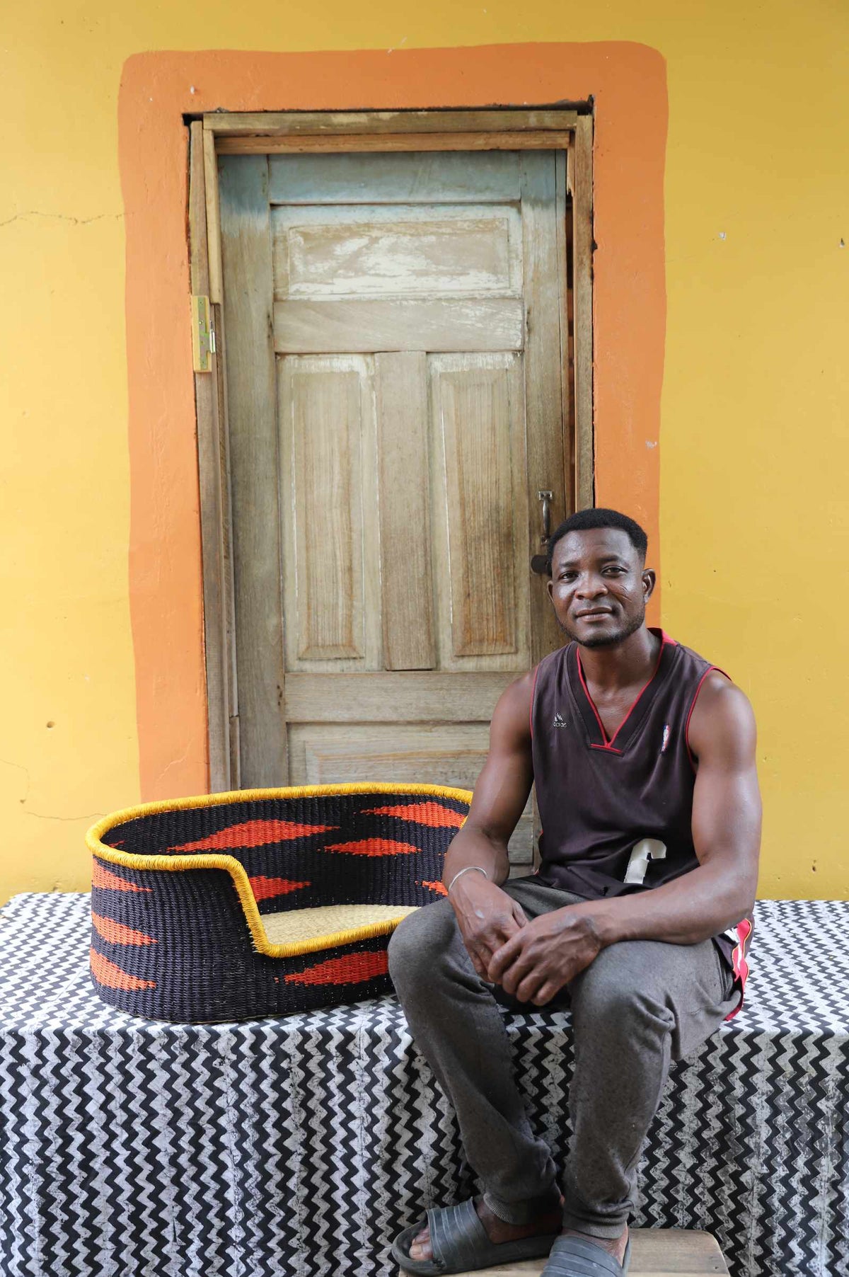 Medium handwoven Bolga pet bed in black with orange flame motif, beside a seated man in a dark vest.