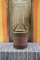 Handwoven open-top Bolga laundry basket in natural straw with black and red woven bands and twin looped handles.