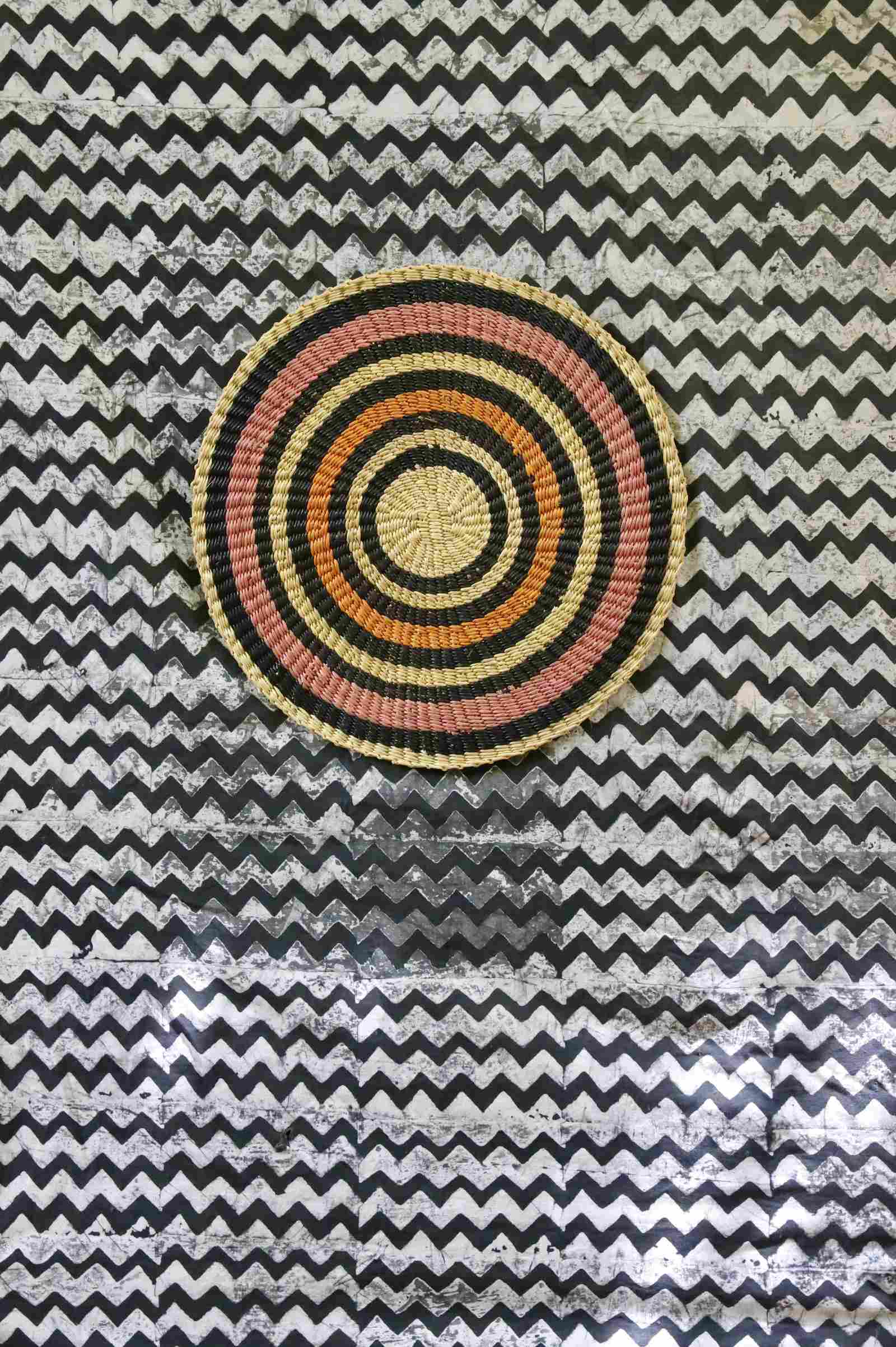 Handwoven Bolga placemat with concentric rings in natural straw, black, orange and pink, on a chevron-print backdrop.