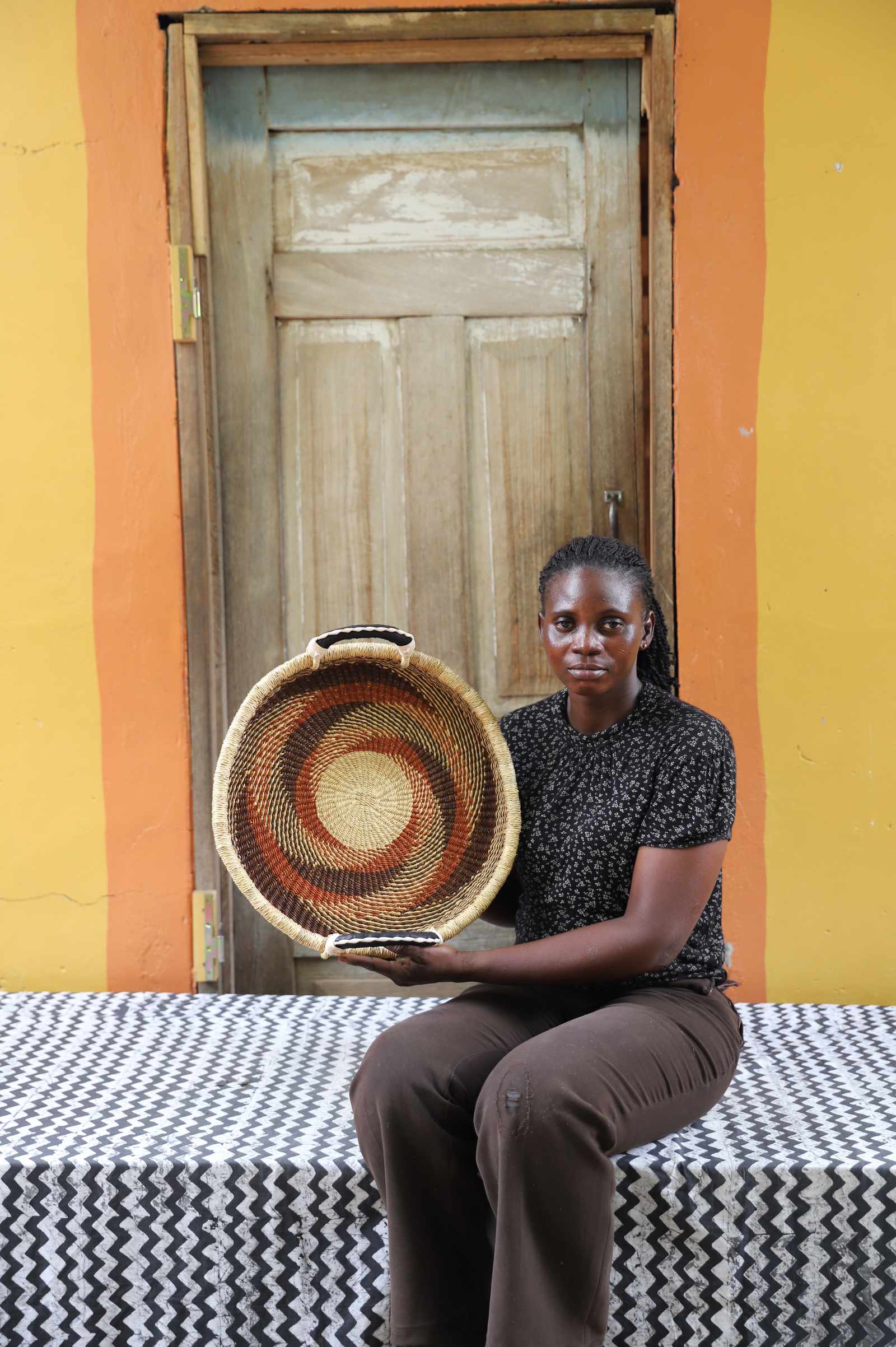 Handwoven Bolga wall tray disc with rust-red pinwheel, held by a seated woman in dark top.