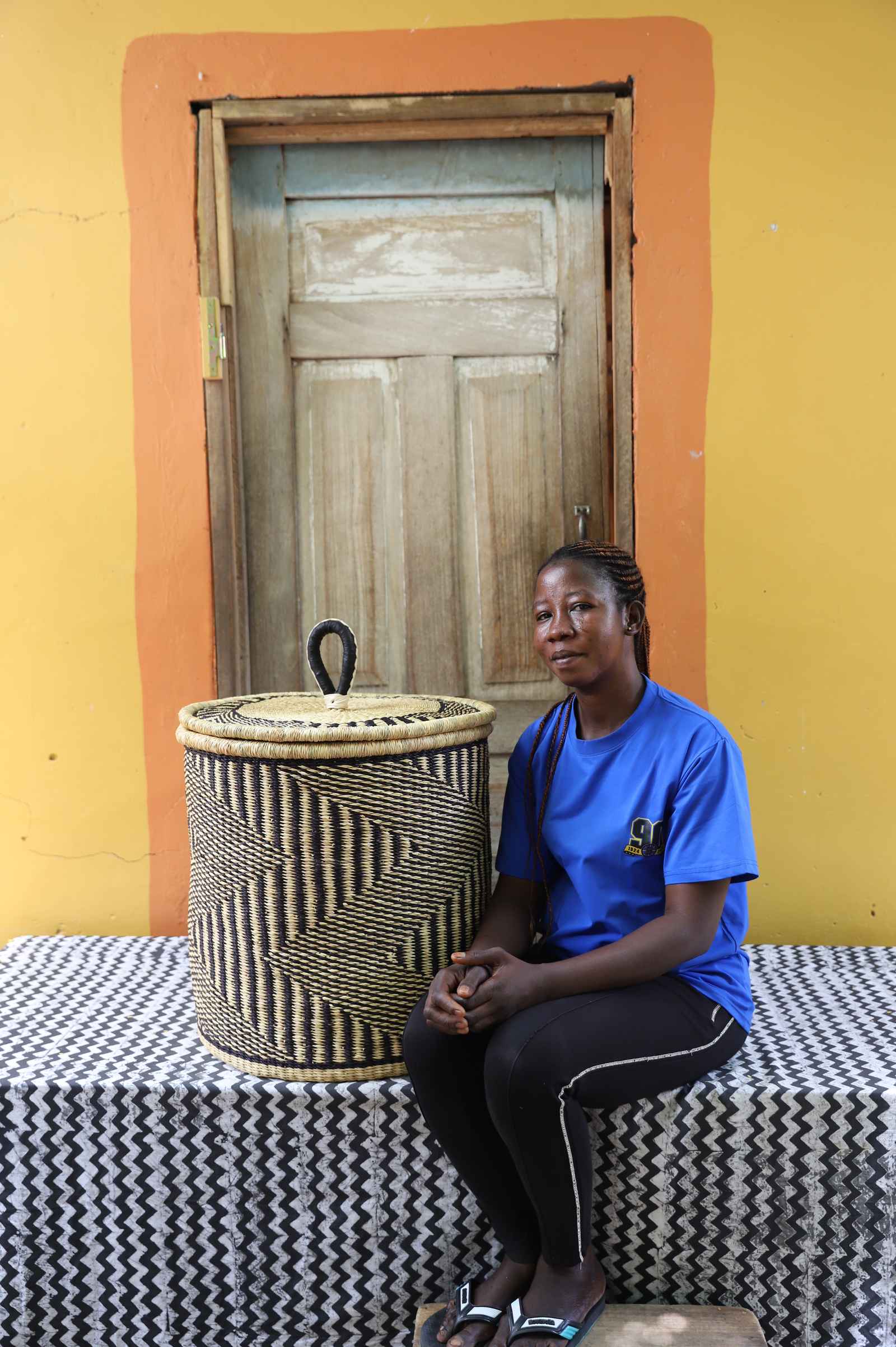Handwoven Bolga laundry basket in natural with dark wave stripes, beside a seated woman in blue.
