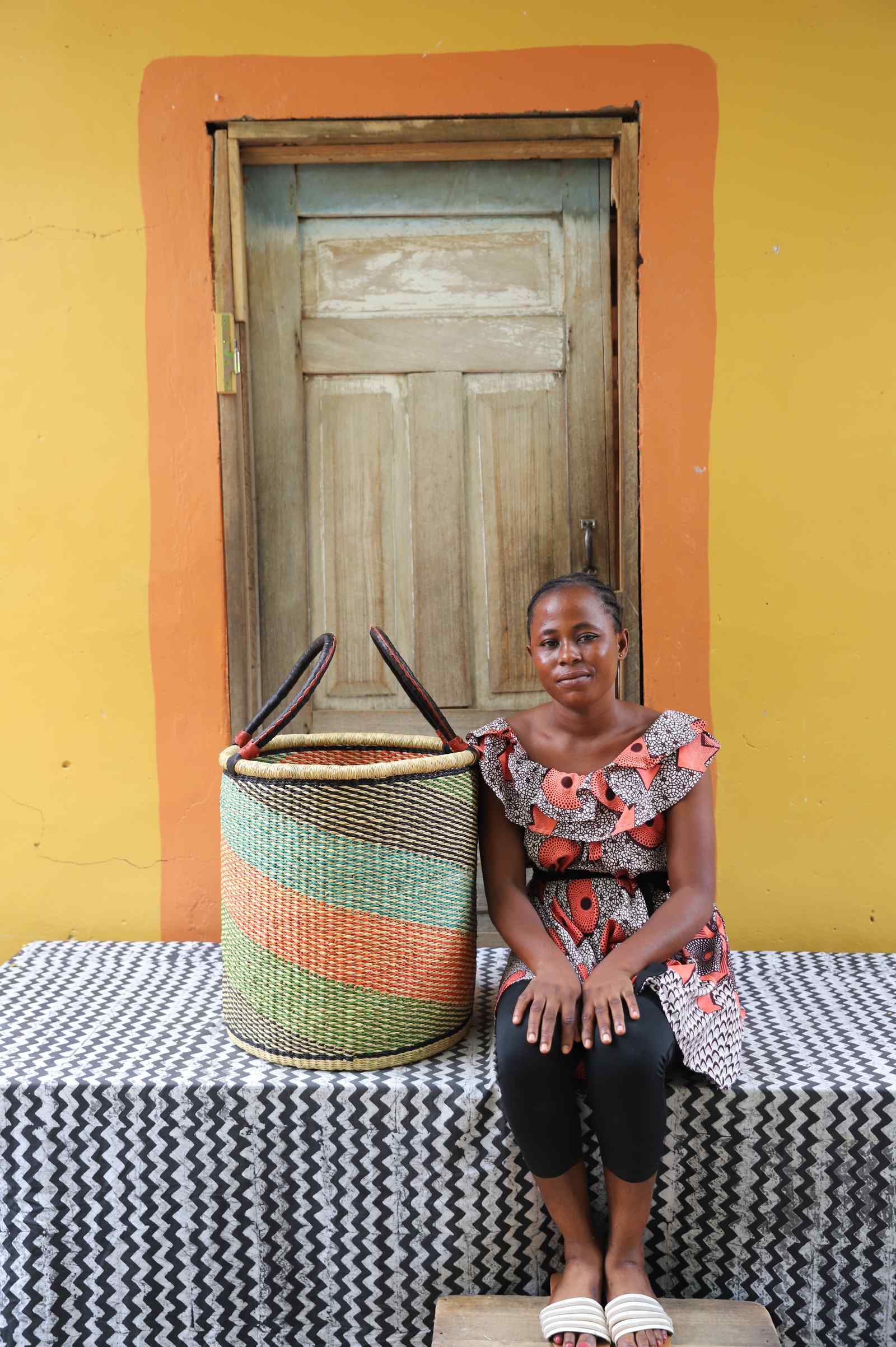 Medium handwoven Bolga laundry basket in green, orange and pink stripes, beside a seated woman in floral.