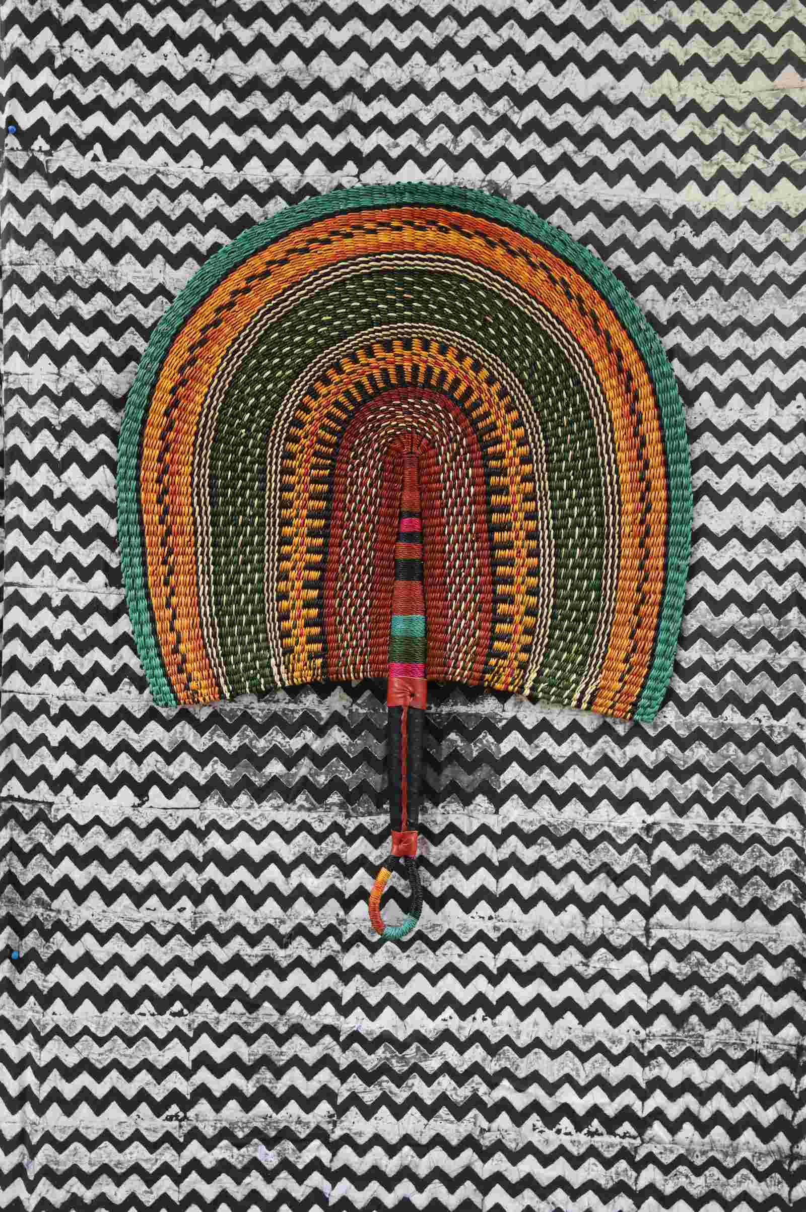 Naba Penga wall fan in natural straw with teal, orange and green concentric bands, laid on a chevron cloth.
