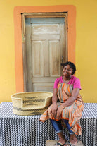 Small handwoven Bolga pet bed in natural straw with subtle black motif, beside a seated woman in pink.
