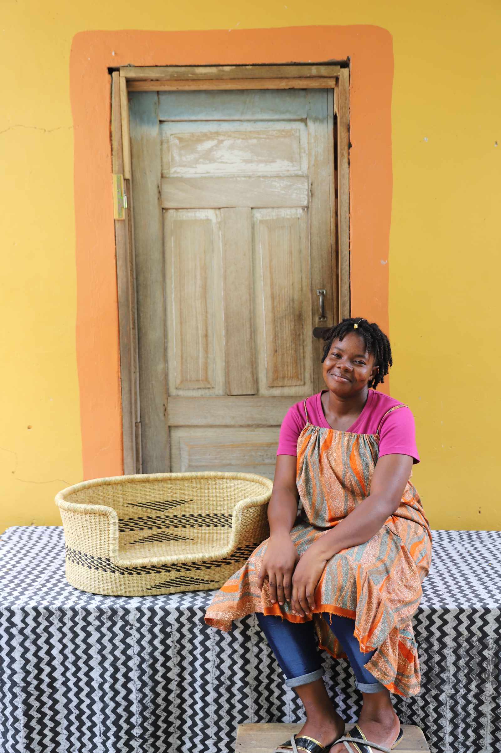 Small handwoven Bolga pet bed in natural straw with subtle black motif, beside a seated woman in pink.