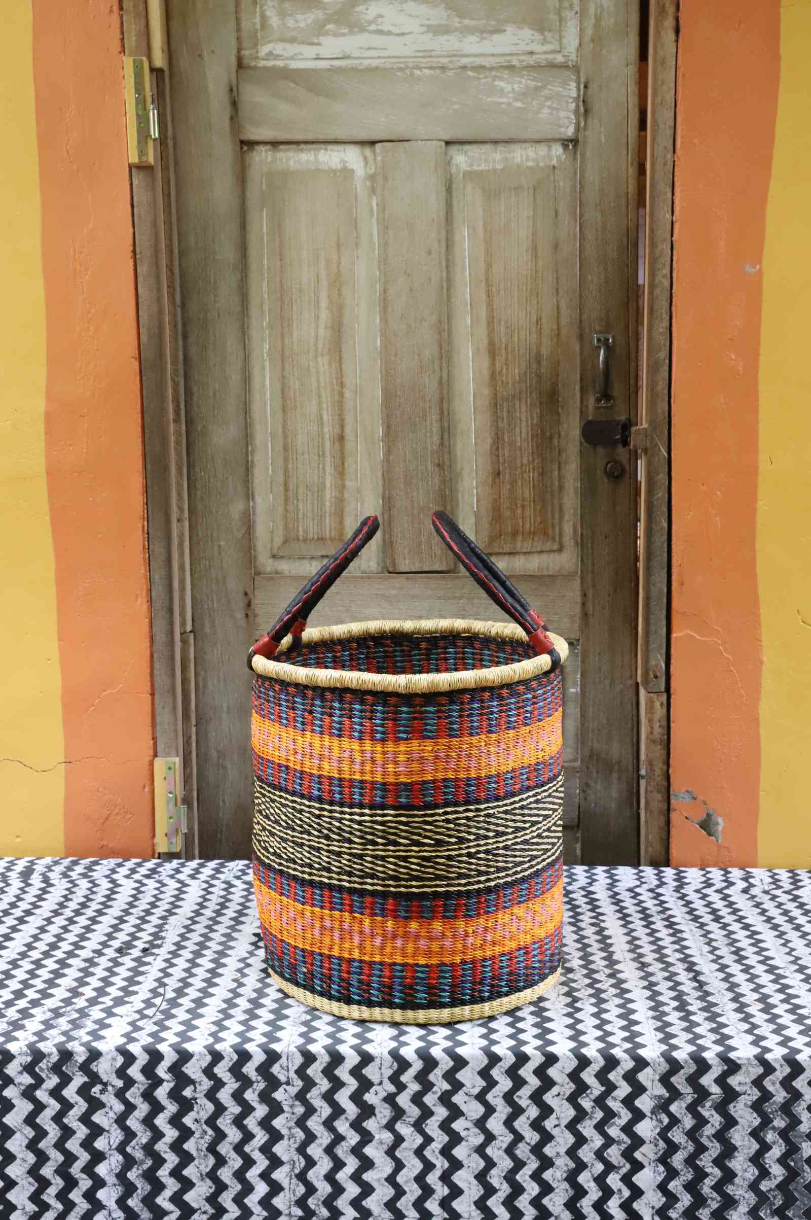 Handwoven open-top Bolga laundry basket in natural straw with orange, yellow and black stripes and twin handles.