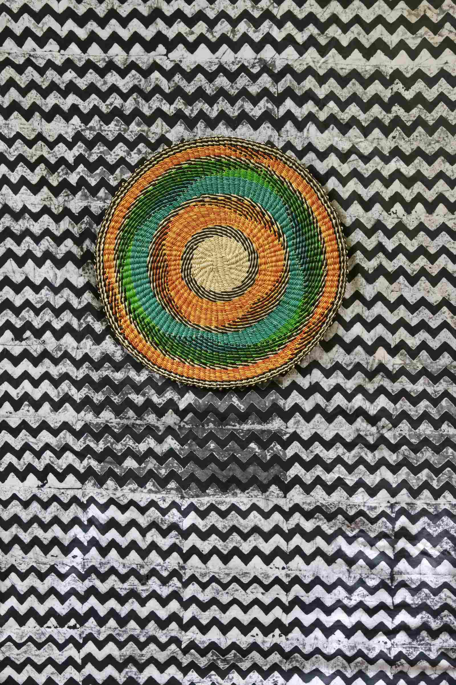 Handwoven Bolga placemat with concentric rings in natural straw, orange, teal and green, on a chevron-print backdrop.