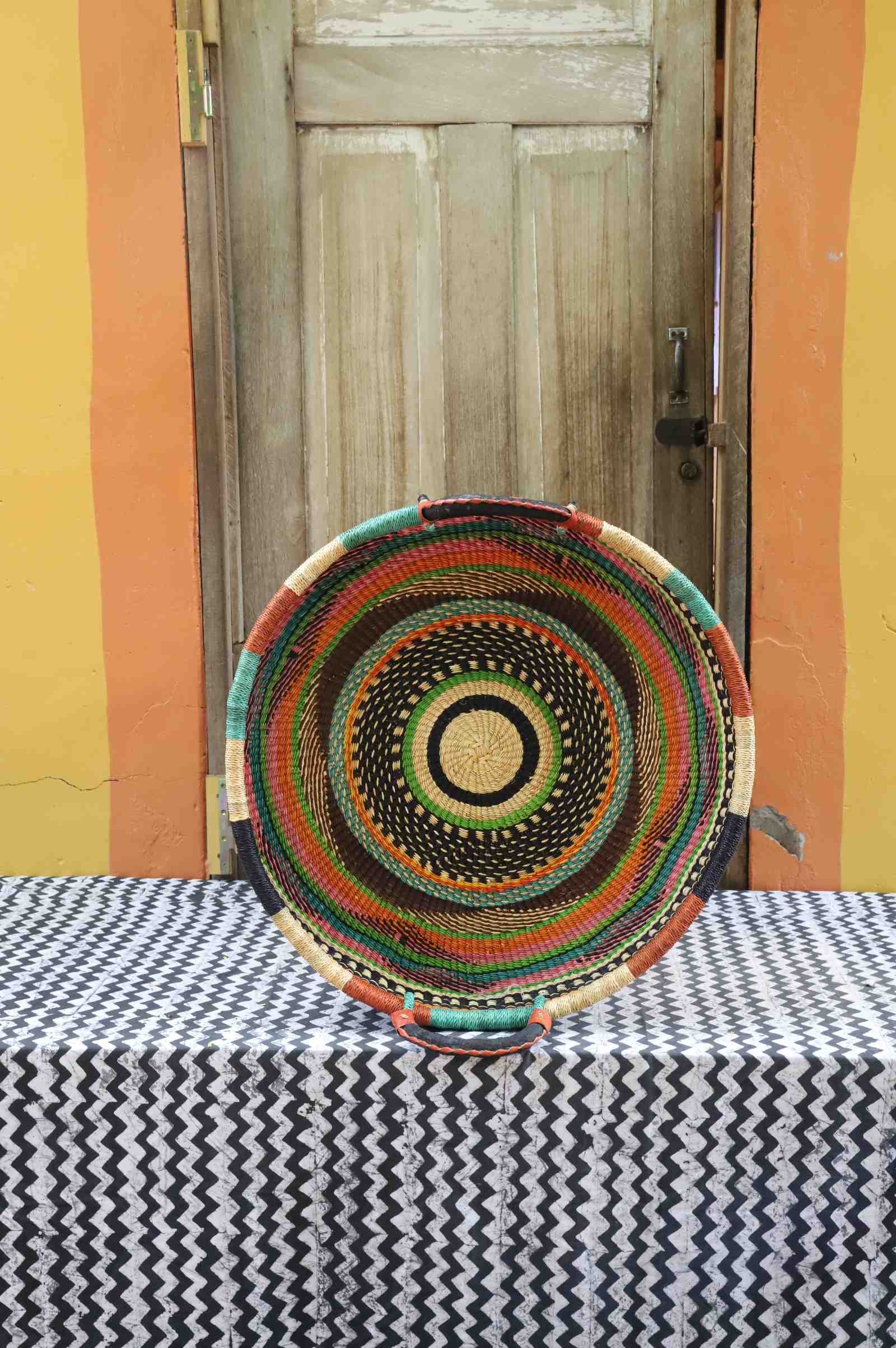 Large handwoven Bolga wall tray disc with orange, green, red and black concentric rings and yellow centre.