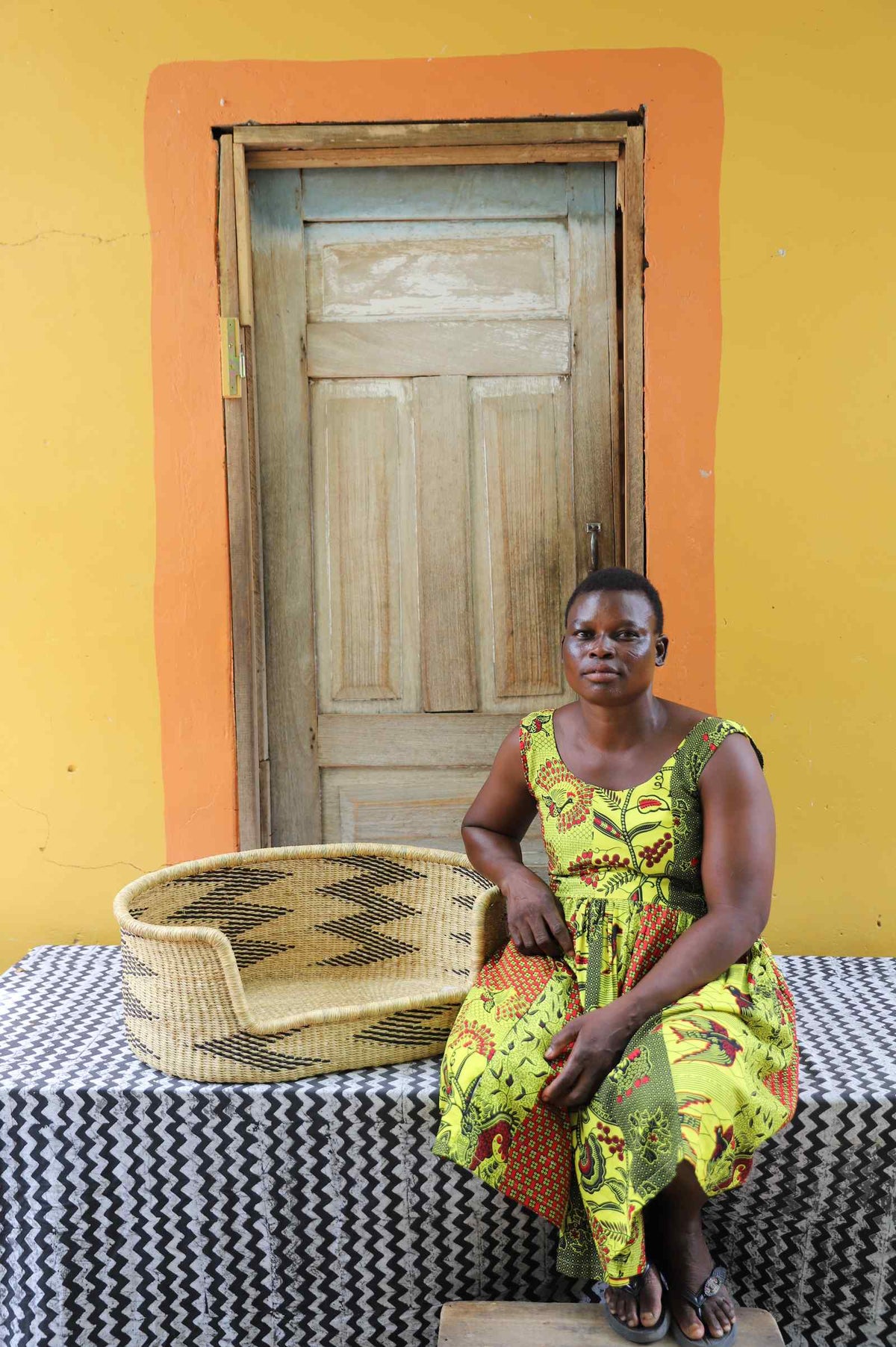 Small handwoven Bolga pet bed in natural straw with black arrow pattern, beside a seated woman in yellow.