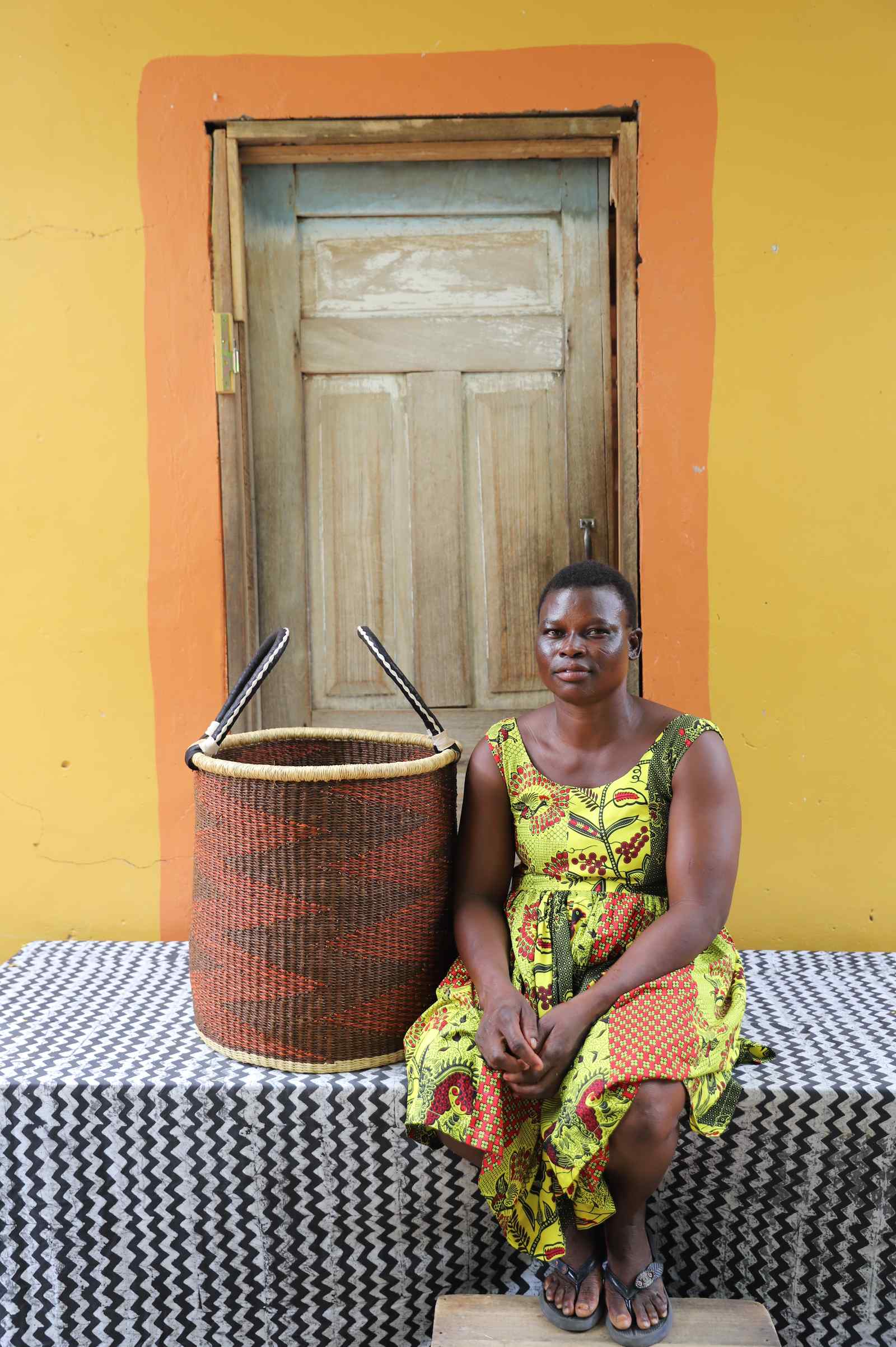 Medium handwoven Bolga laundry basket in rust-orange with dark zigzag, beside a seated woman in floral.