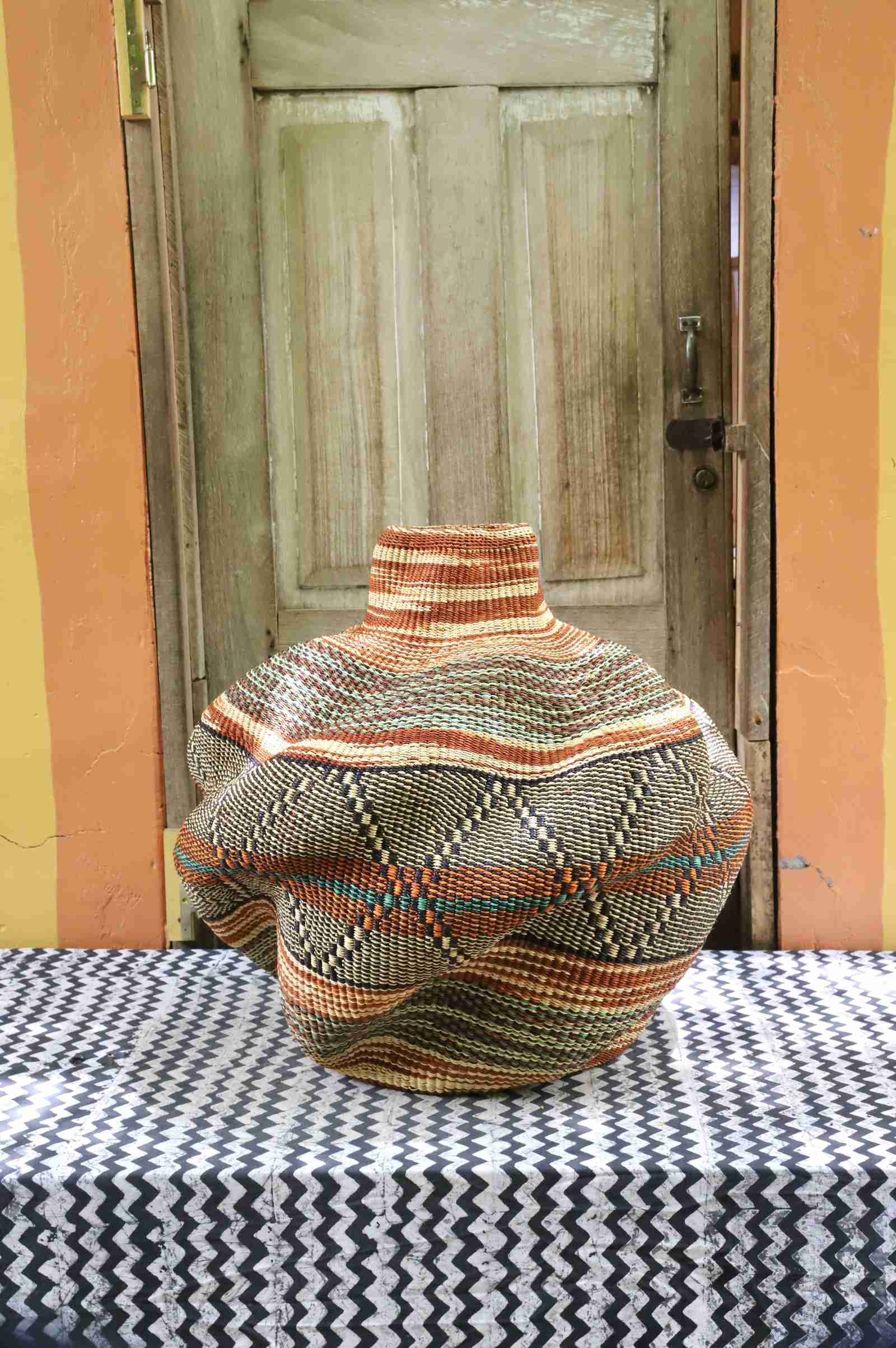 Rear view of a handwoven African art basket showing its sculptural lobed form and earth-tone stripes, on a chevron surface.