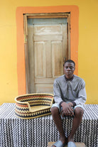 Small handwoven Bolga pet bed in natural straw with black zigzag, beside a seated man in grey.