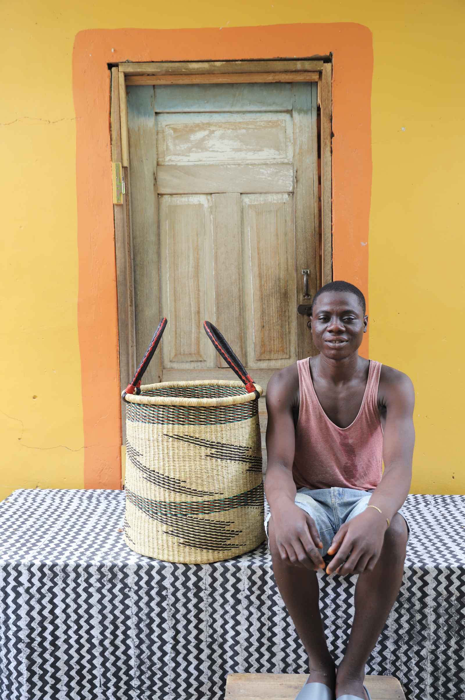 Medium handwoven Bolga laundry basket in natural with scattered black waves, beside a seated man in coral.