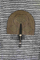 Naba Penga wall fan in natural straw with a dark geometric diamond weave, laid on a chevron cloth.