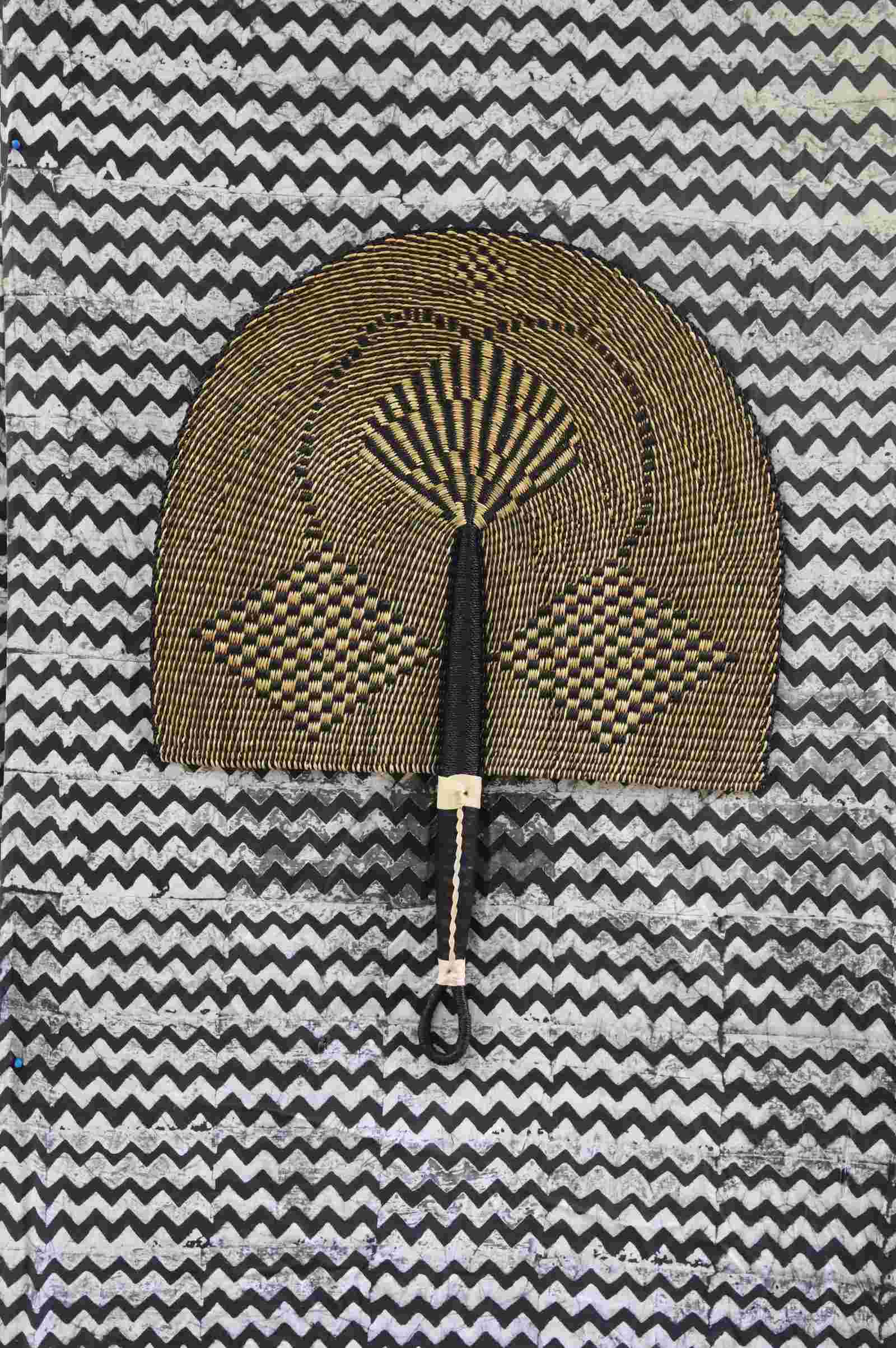 Naba Penga wall fan in natural straw with a dark geometric diamond weave, laid on a chevron cloth.