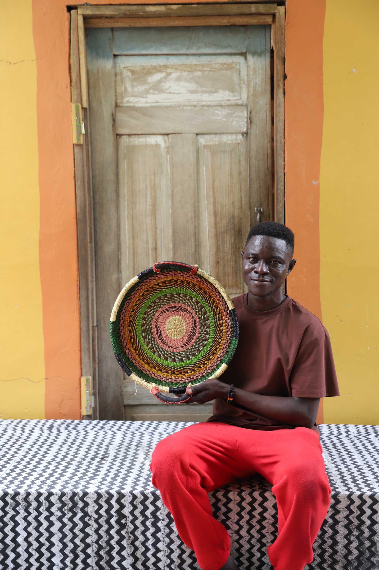 Handwoven Bolga wall tray disc with red and green bands, held by a seated man in a brown top.