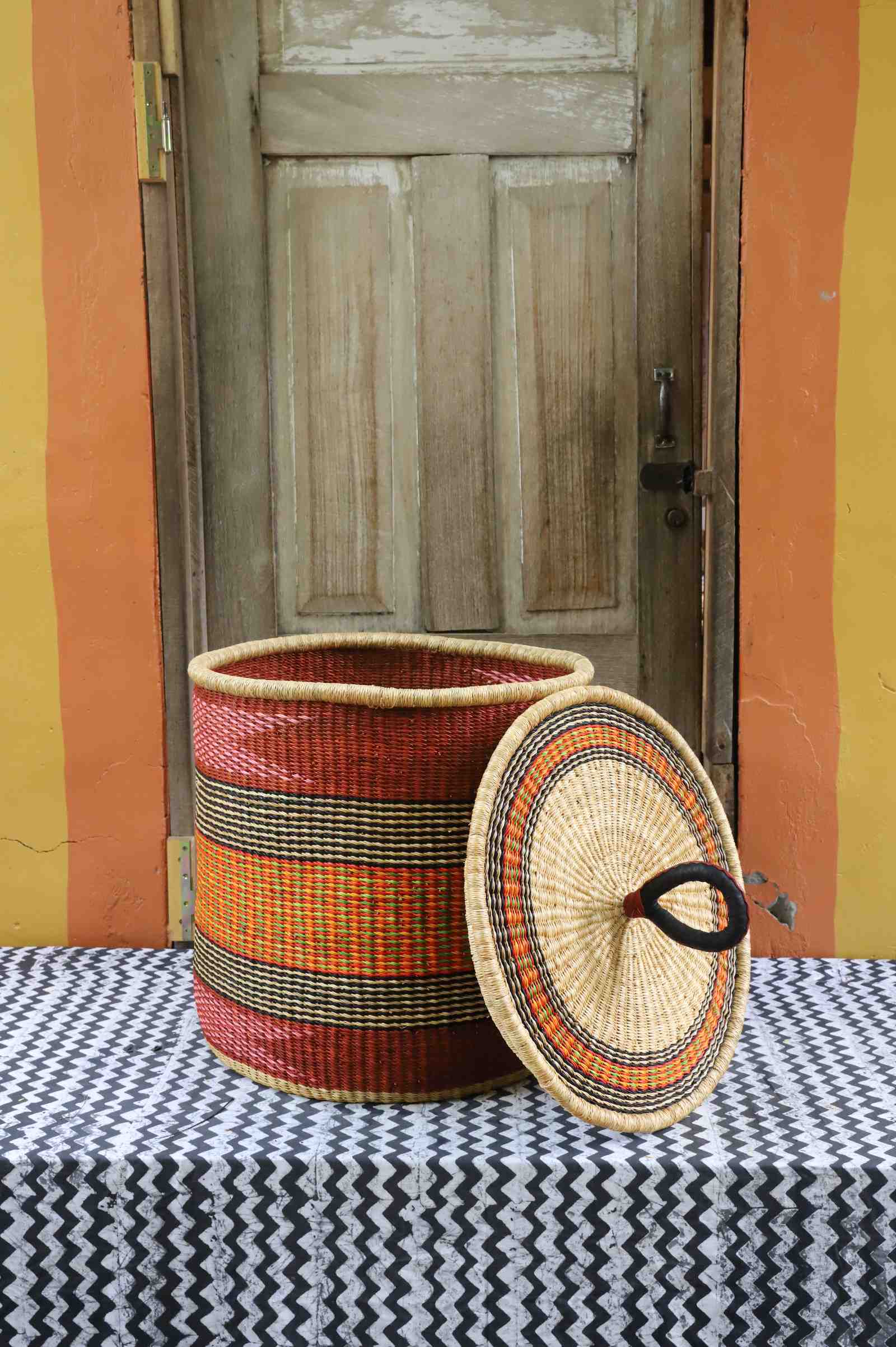 Handwoven Bolga laundry basket with lid removed, deep red body with orange and yellow banded stripes.