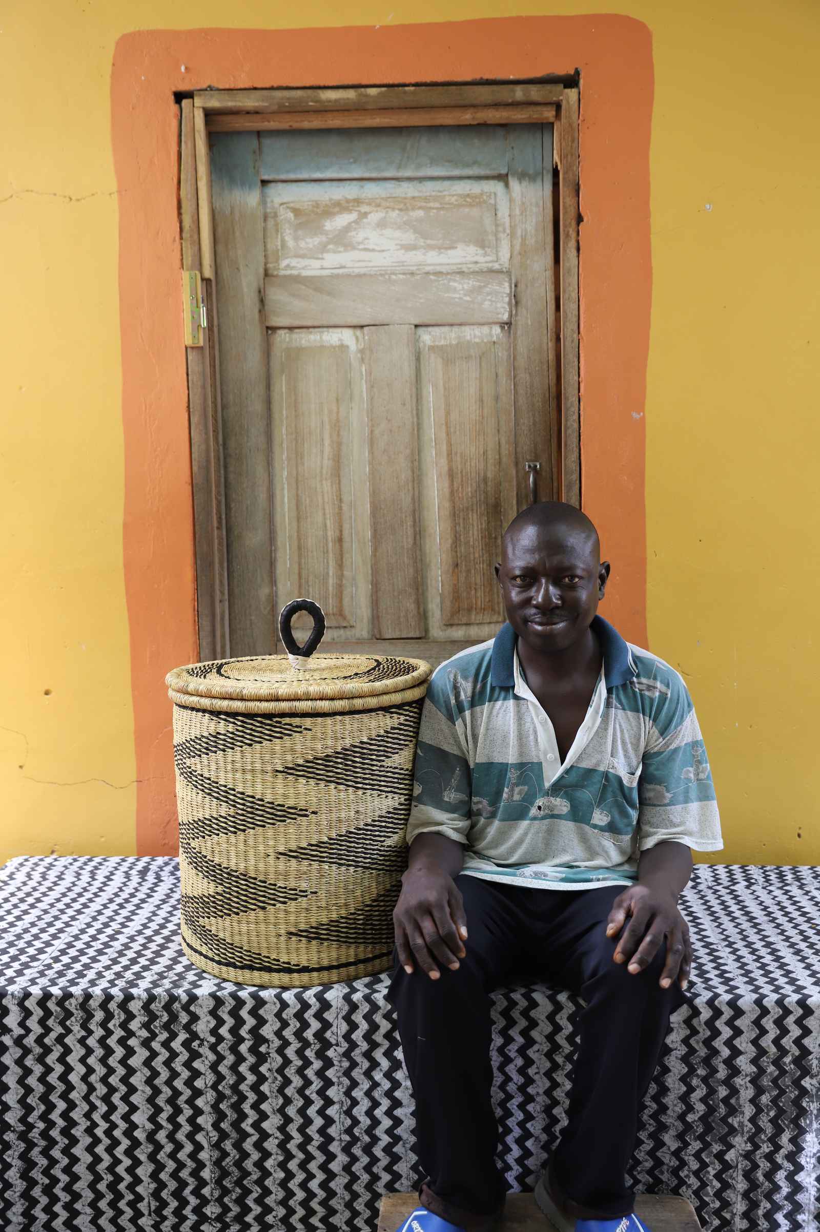 Handwoven Bolga laundry basket in natural straw with black wave motif, beside a seated man in stripes.