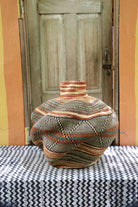 Handwoven African art basket in sculptural lobed vase form with multicoloured earth-tone stripes, on a chevron surface.