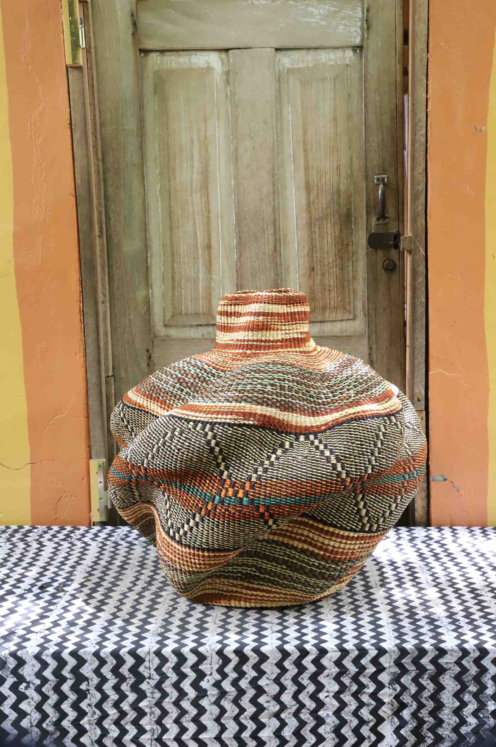 Handwoven African art basket in sculptural lobed vase form with multicoloured earth-tone stripes, on a chevron surface.