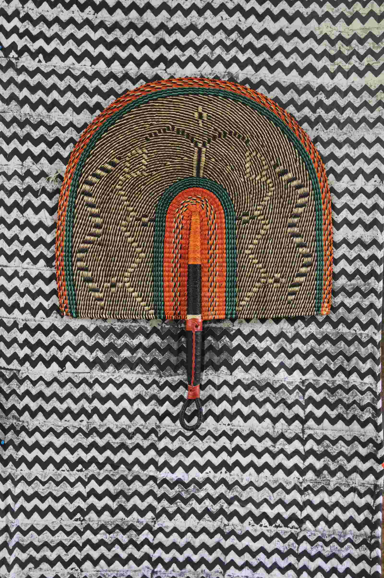 Naba Penga wall fan in natural straw with an orange and green banded border, laid on a chevron cloth.