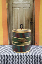 Handwoven Bolga laundry basket with coiled rim and lid in natural straw with a dark green band and arrow motif.