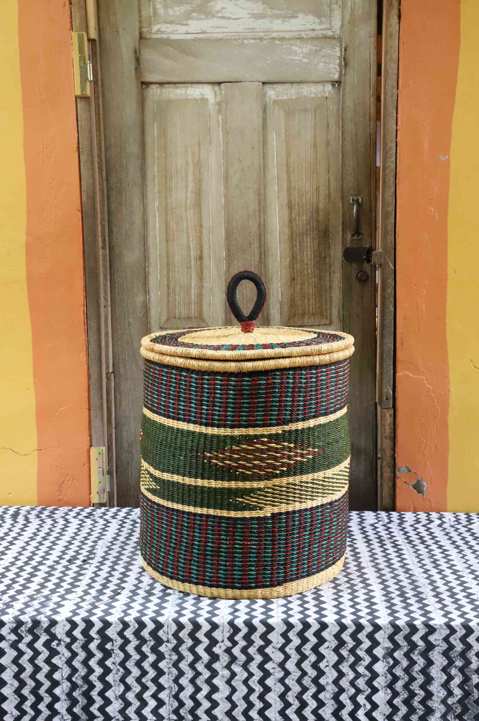 Handwoven Bolga laundry basket with coiled rim and lid in natural straw with a dark green band and arrow motif.