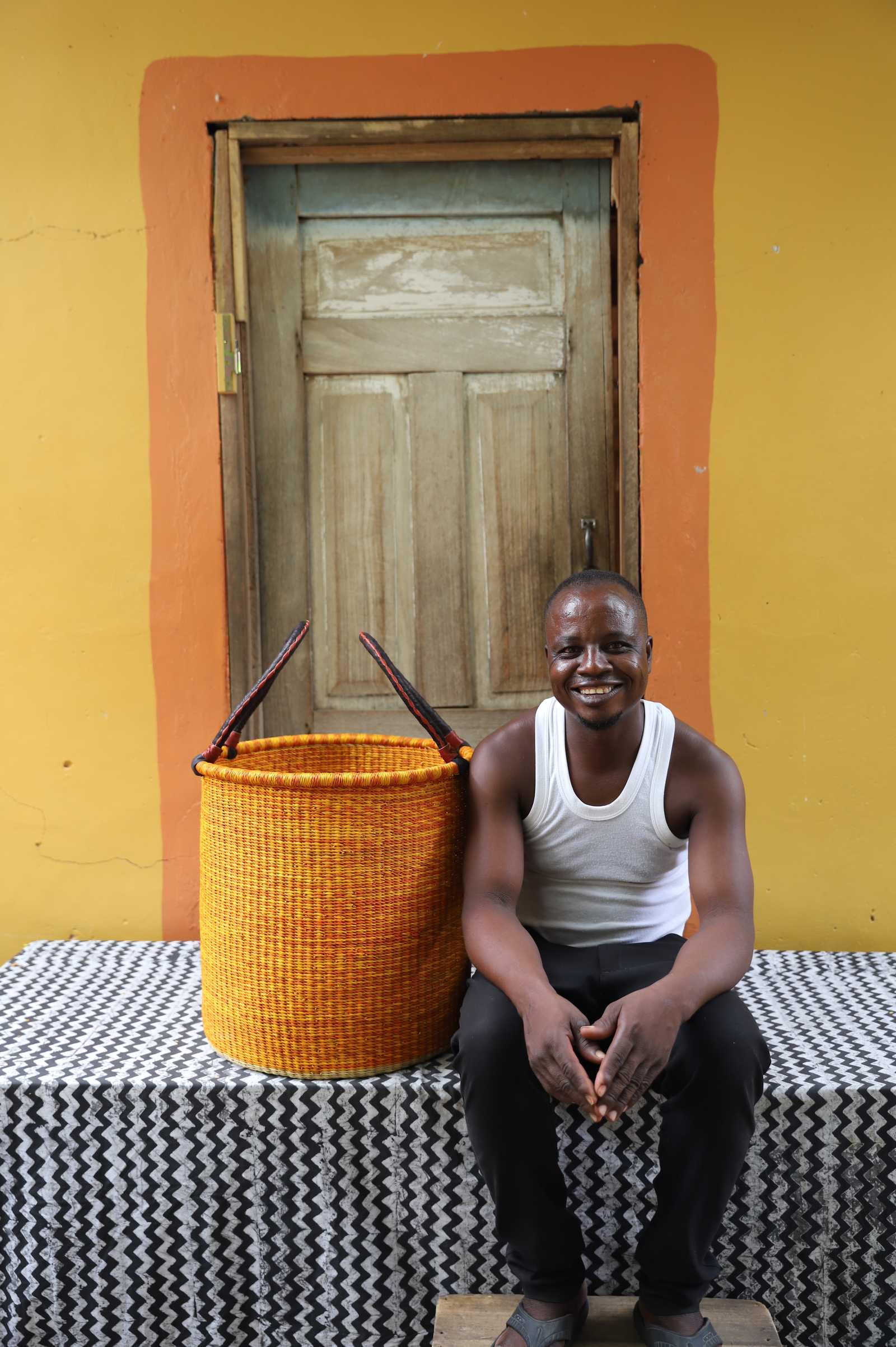 Medium handwoven Bolga laundry basket in dyed bold orange, beside a seated man in a white vest.