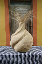 Handwoven art basket in natural straw with sculptural twisted body and dried grass stems sprouting from the top.