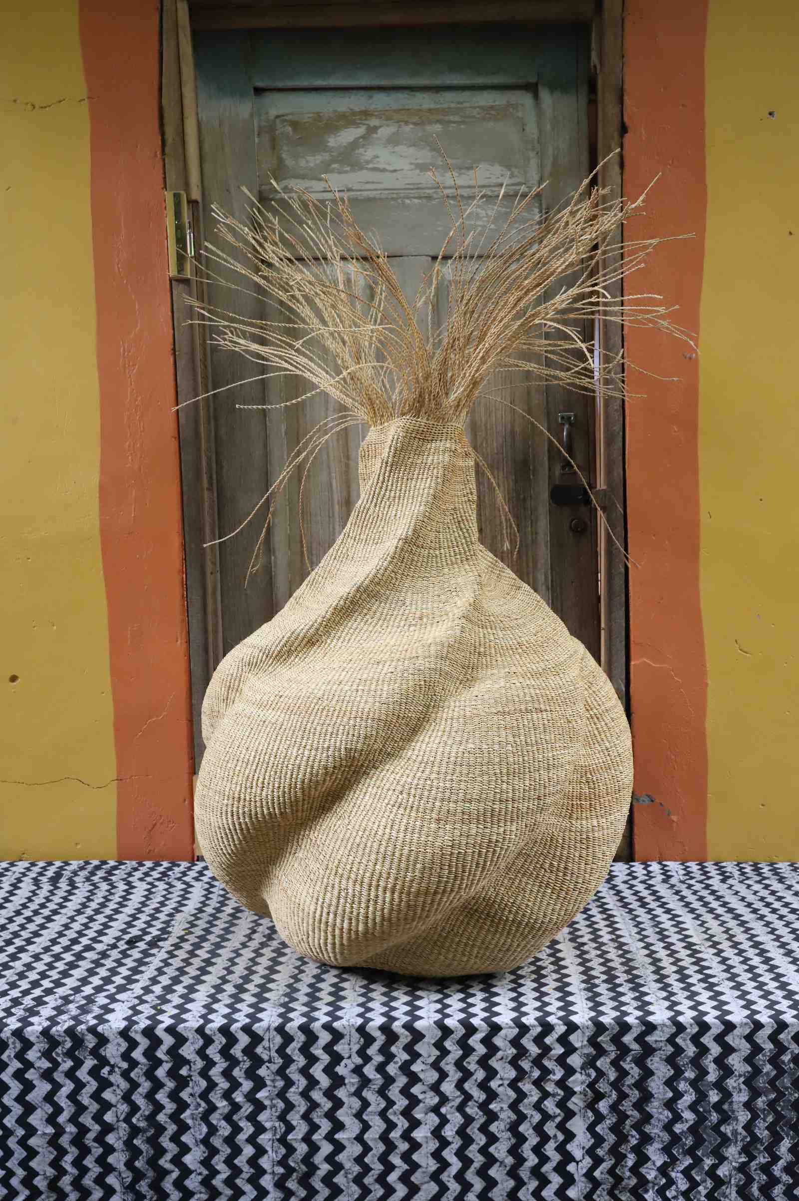 Handwoven art basket in natural straw with sculptural twisted body and dried grass stems sprouting from the top.