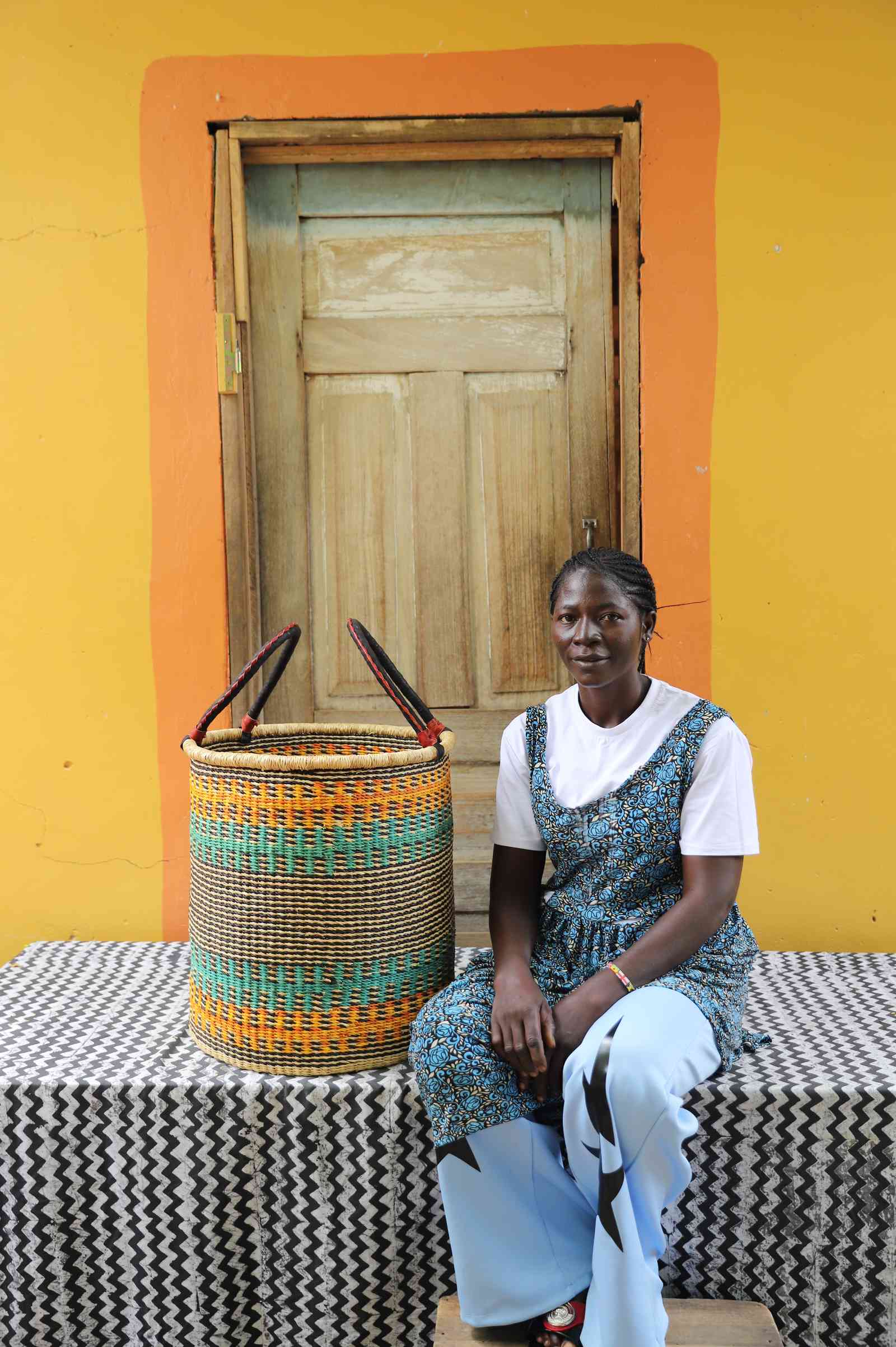 Medium handwoven Bolga laundry basket in natural with orange and teal bands, beside a seated woman in blue.