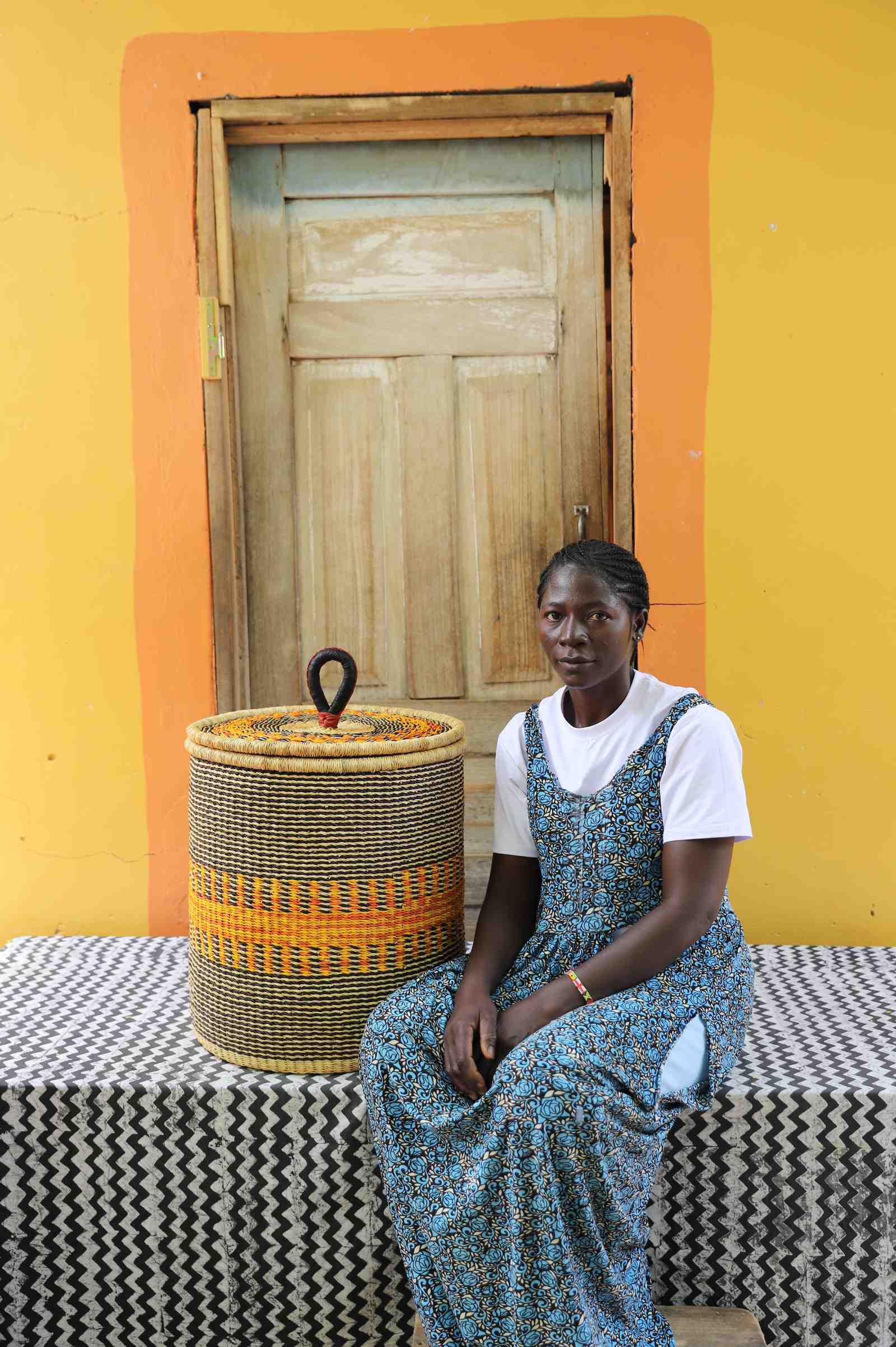 Handwoven Bolga laundry basket in natural straw with orange central band, beside a seated woman in floral.