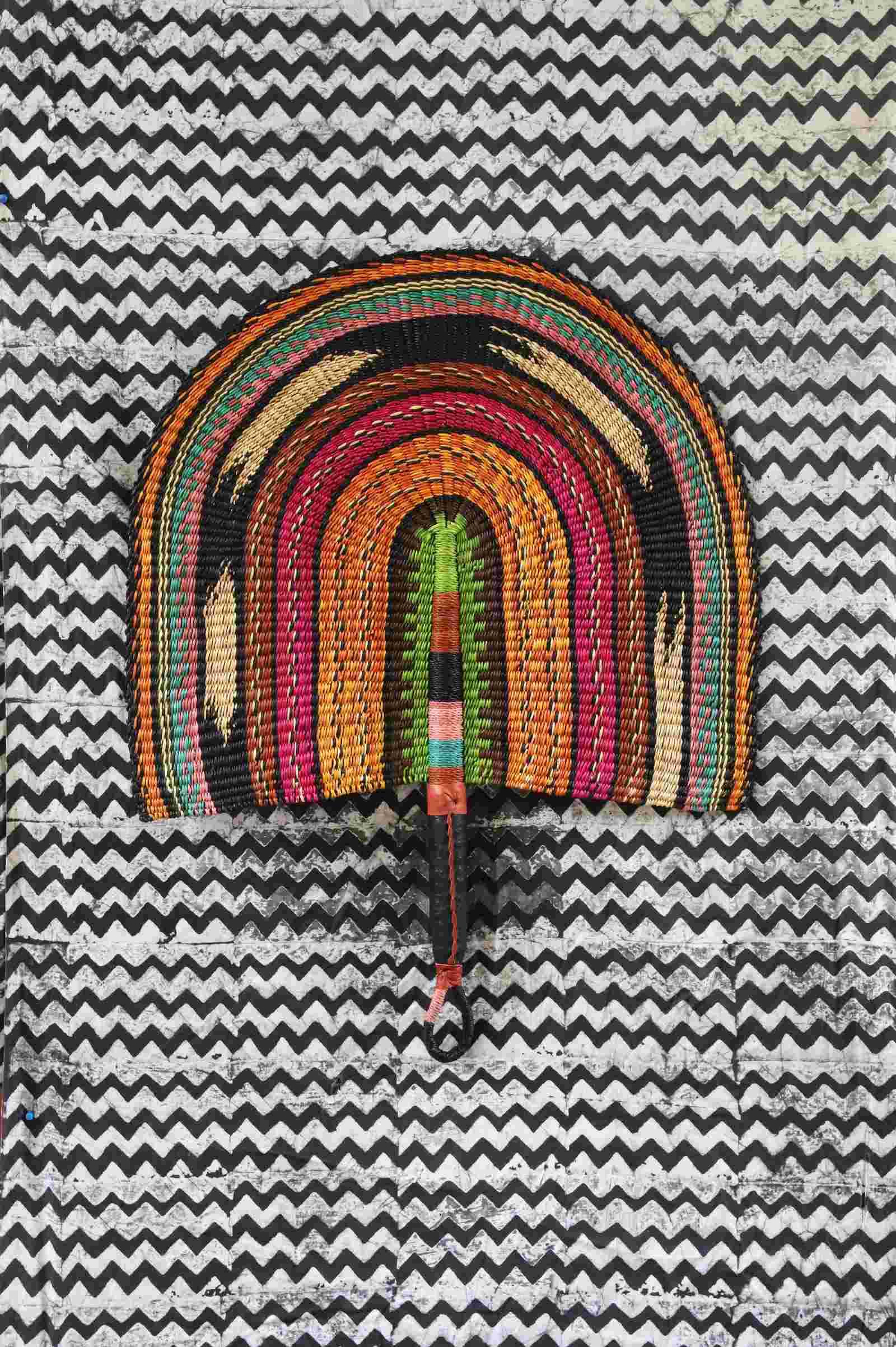 Naba Penga wall fan in natural straw with rust, green, pink and black rainbow arches, laid on a chevron cloth.