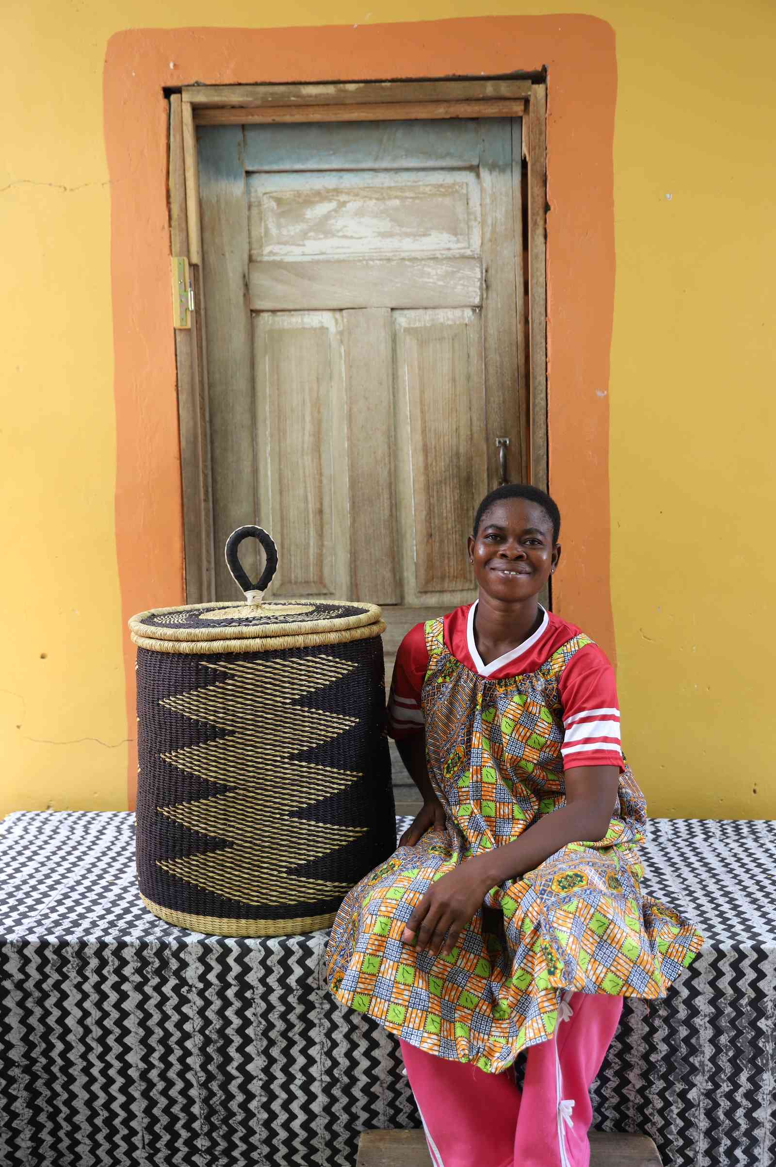 Handwoven Bolga laundry basket in natural with black zigzag band, beside a seated woman in a patterned dress.