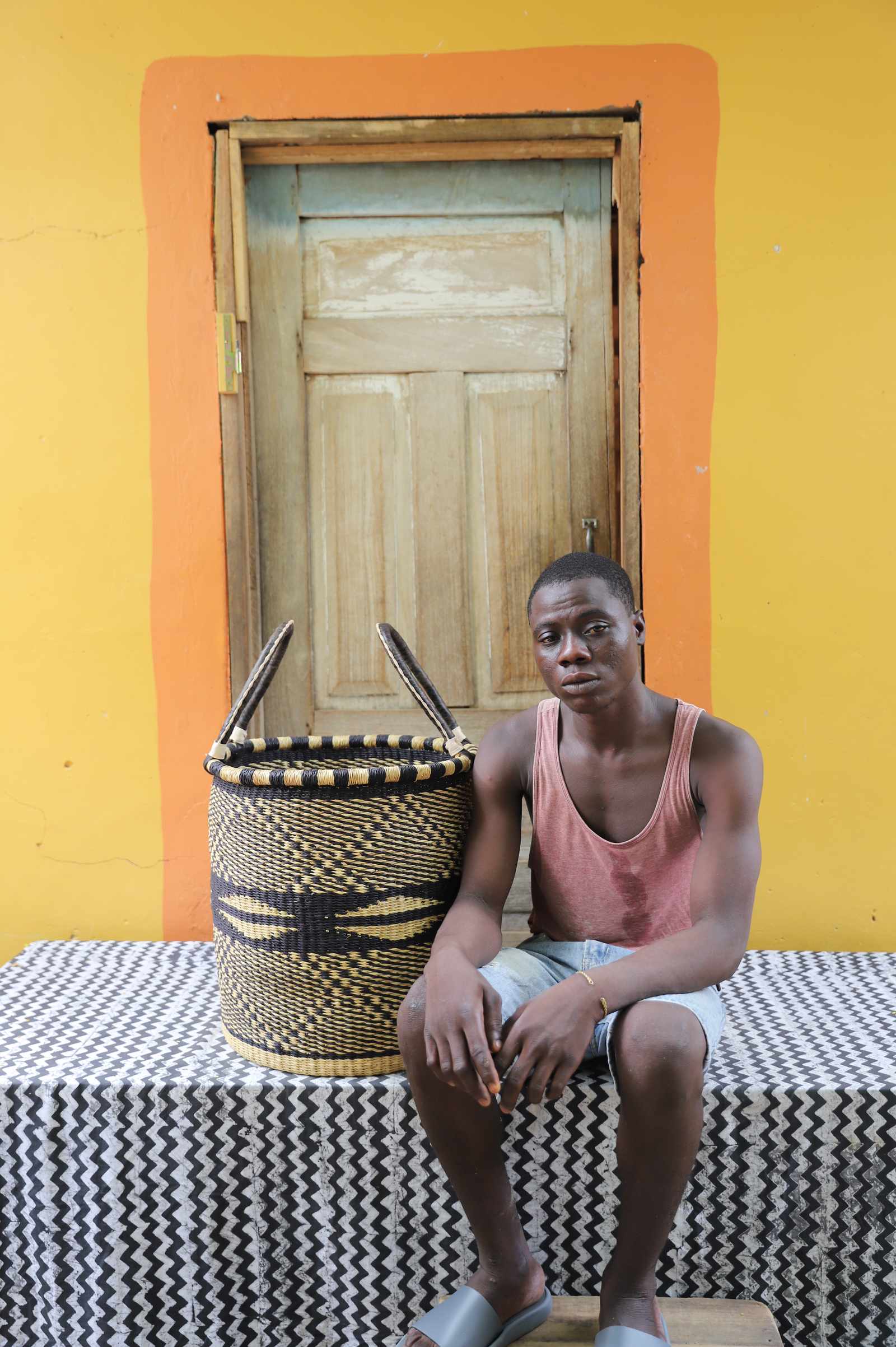 Medium handwoven Bolga laundry basket in natural with black wave motifs, beside a seated man in coral.