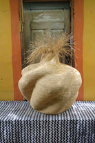 Side view of a handwoven natural straw art basket showing its organic lobed form and dried grass stems, on a chevron surface.