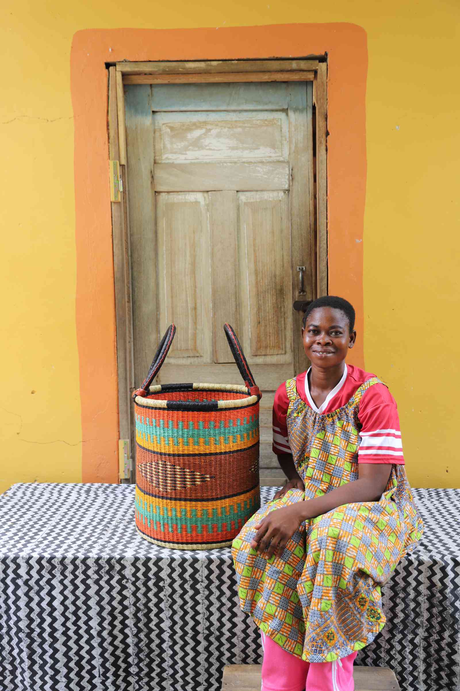 Small handwoven Bolga laundry basket in dyed orange with green and red bands, beside a seated woman.