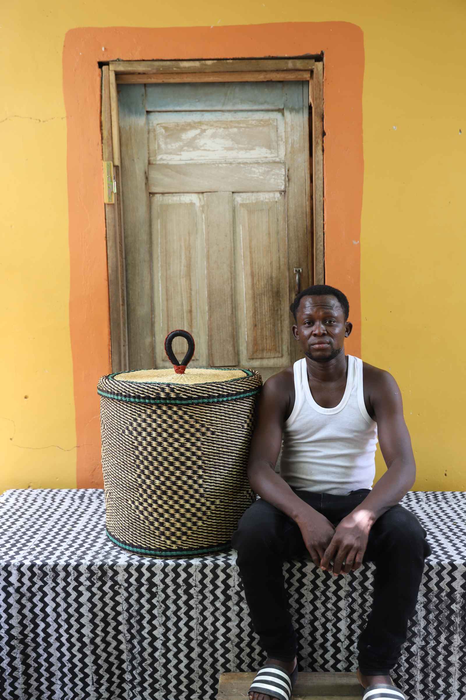 Handwoven Bolga laundry basket in natural straw with black checker pattern, beside a seated man in a white vest.