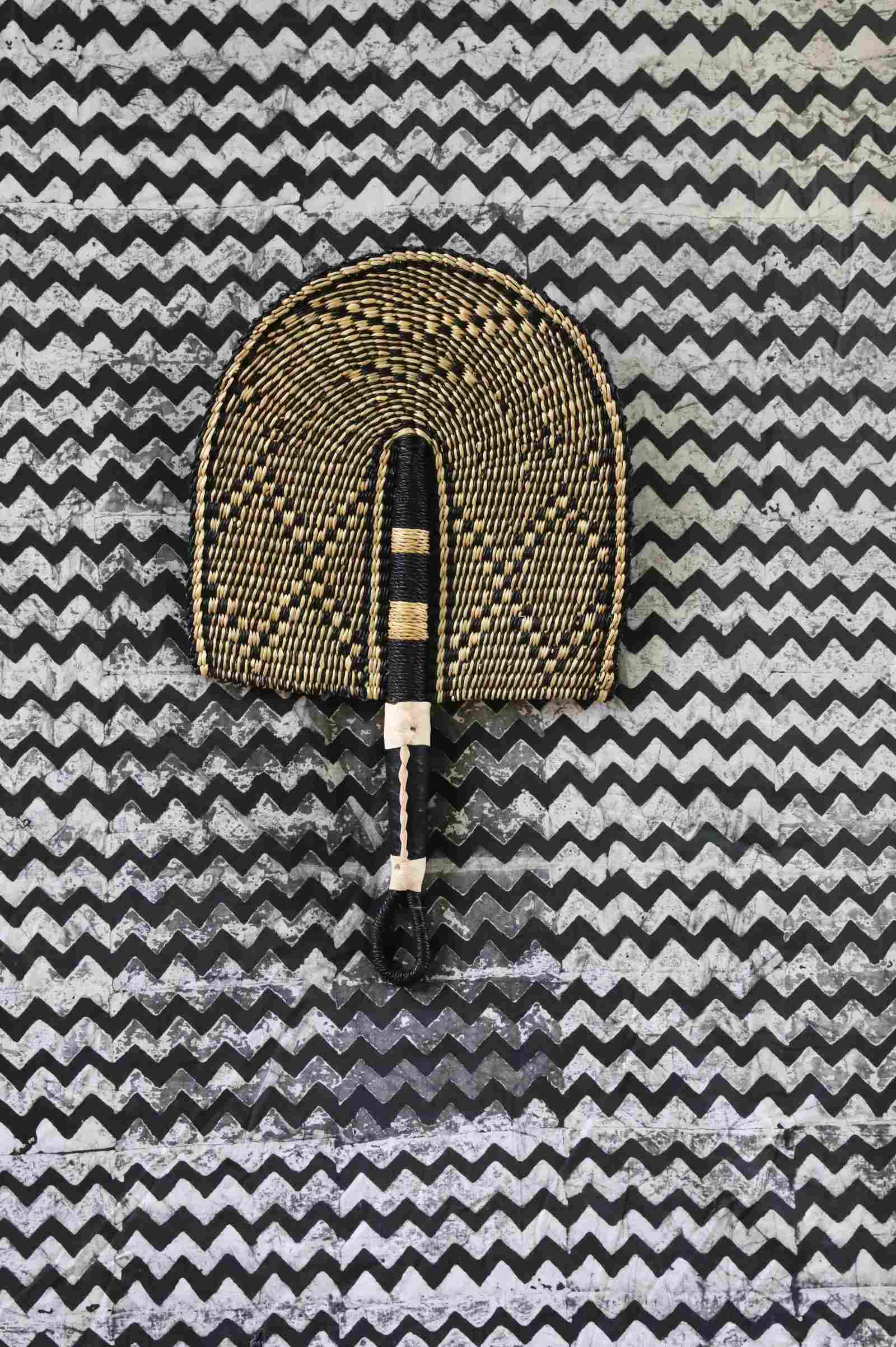 Bolga hand fan in natural straw with a dark chevron pattern and beaded handle, laid on a chevron cloth.