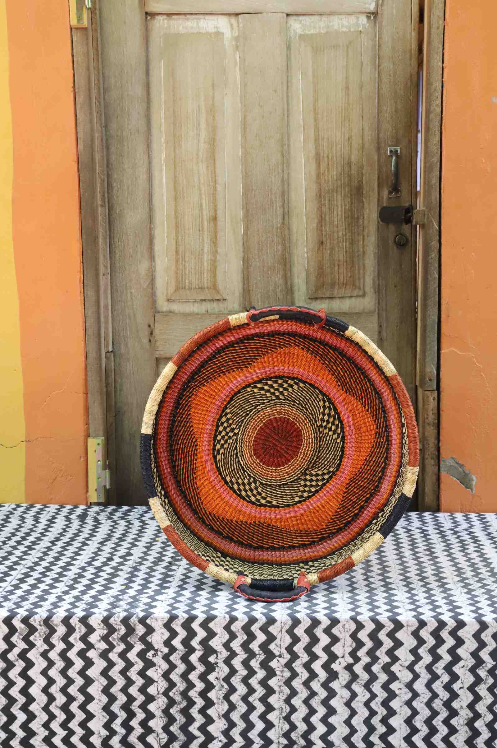Handwoven Bolga wall tray disc in dyed rust-orange with concentric black ring pattern.