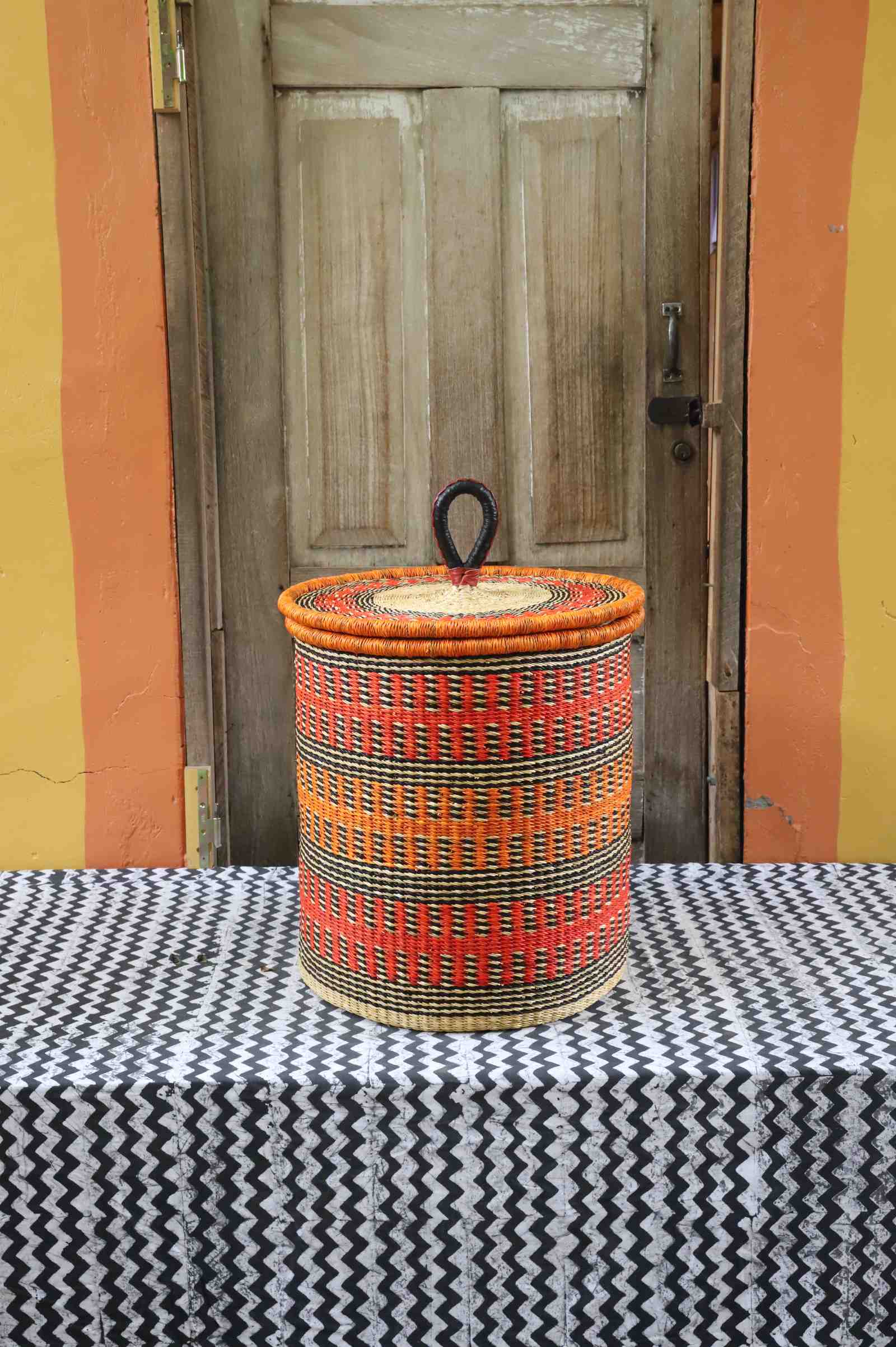 Coiled-rim lidded laundry basket in orange and black fine-striped weave, styled on a chevron cloth by a wooden door.