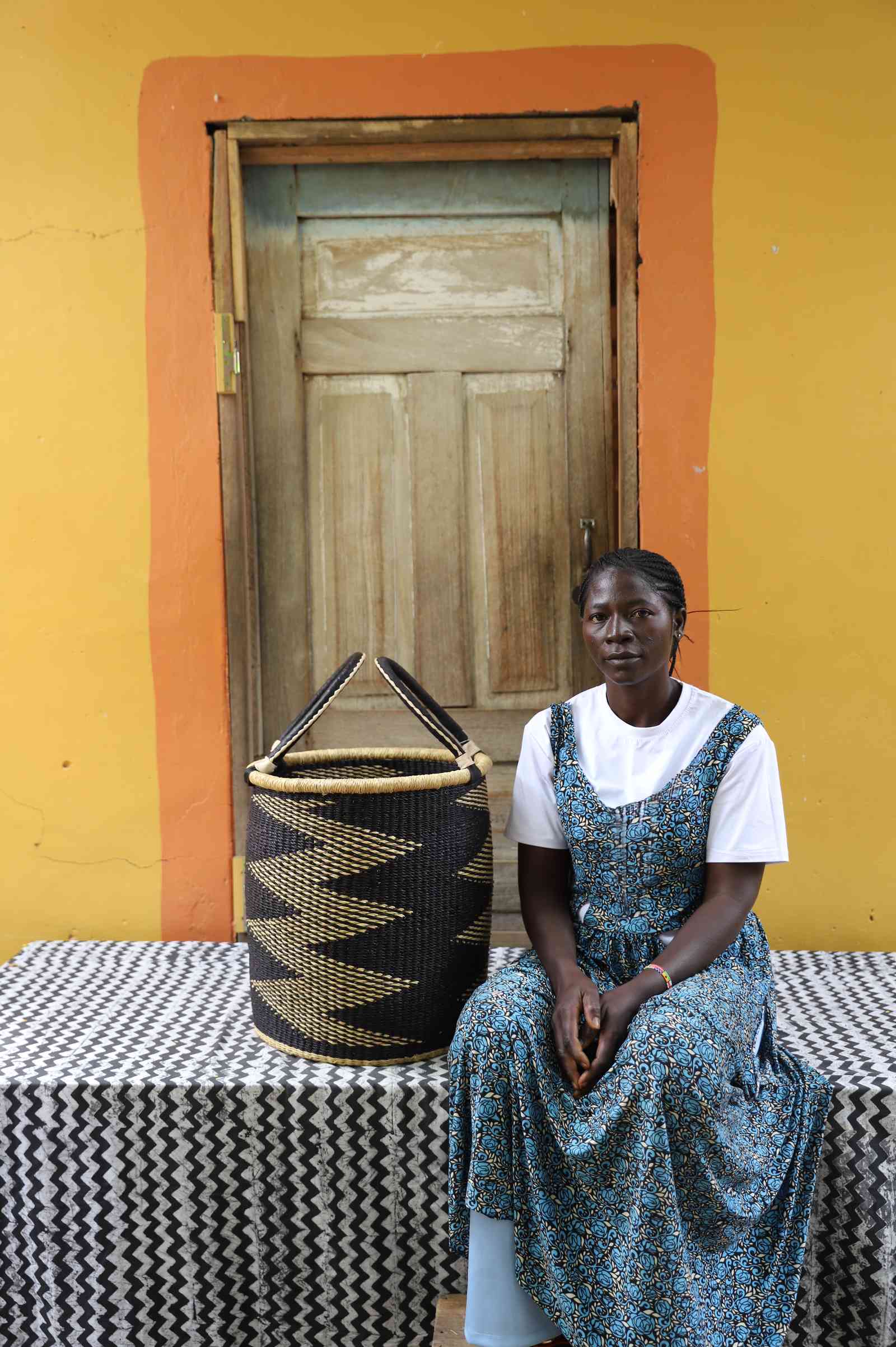 Small handwoven Bolga laundry basket in natural with bold black zigzag, beside a seated woman in green.