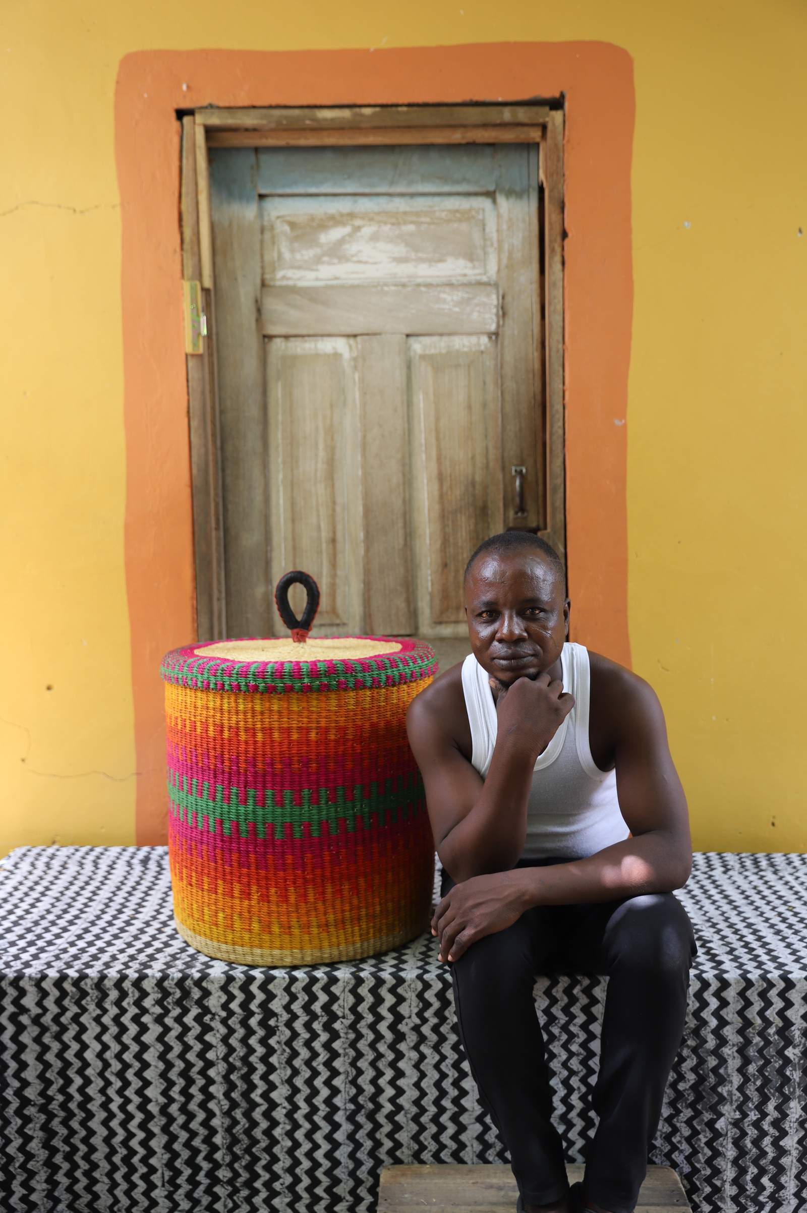 Handwoven Bolga laundry basket in fine multicolour stripes, beside a seated man in a white vest.