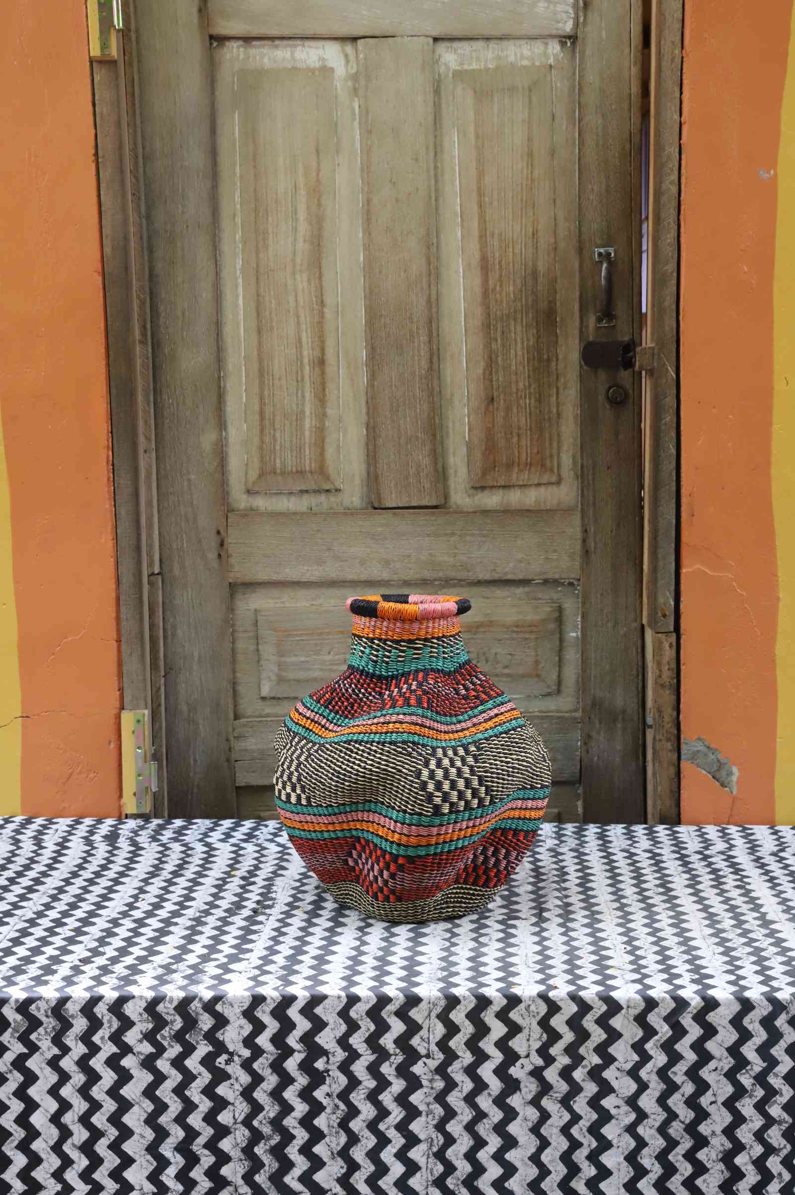 Handwoven Bolga art basket in an urn shape, natural straw with teal, red, green and yellow banded body.
