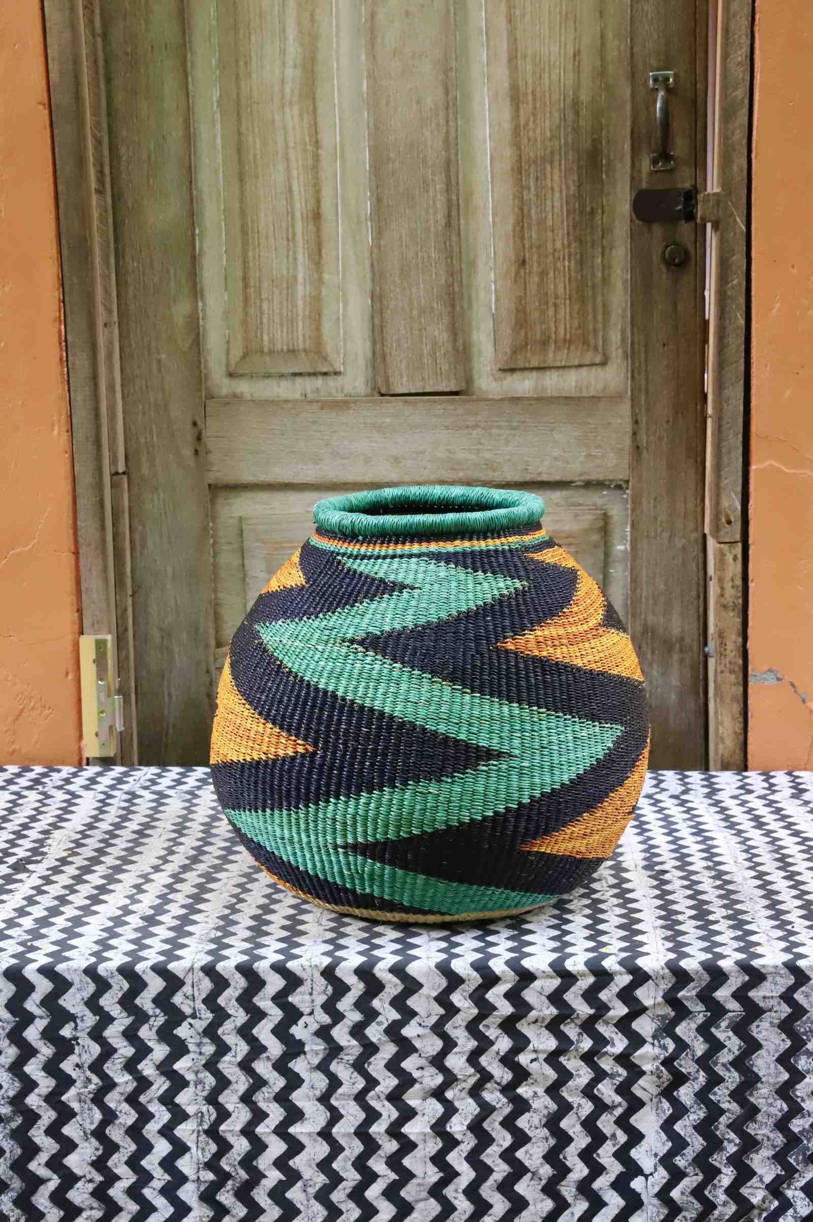 Handwoven African art basket with bold diagonal zigzag pattern in teal, black and amber, on a chevron surface.
