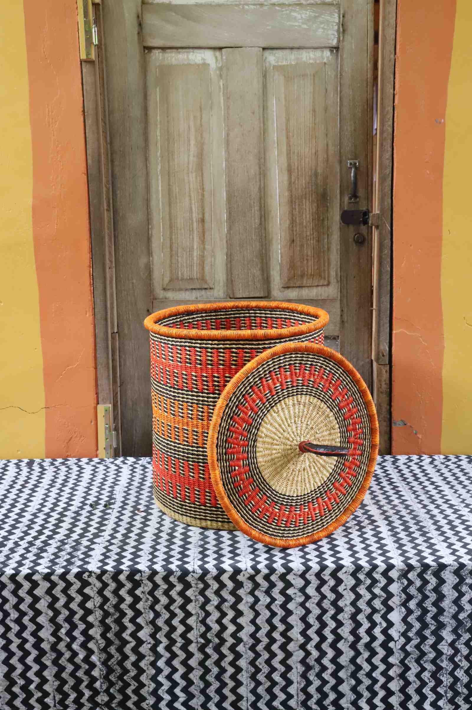Coiled-rim laundry basket in orange and black fine-striped weave, lid resting alongside on a chevron cloth.