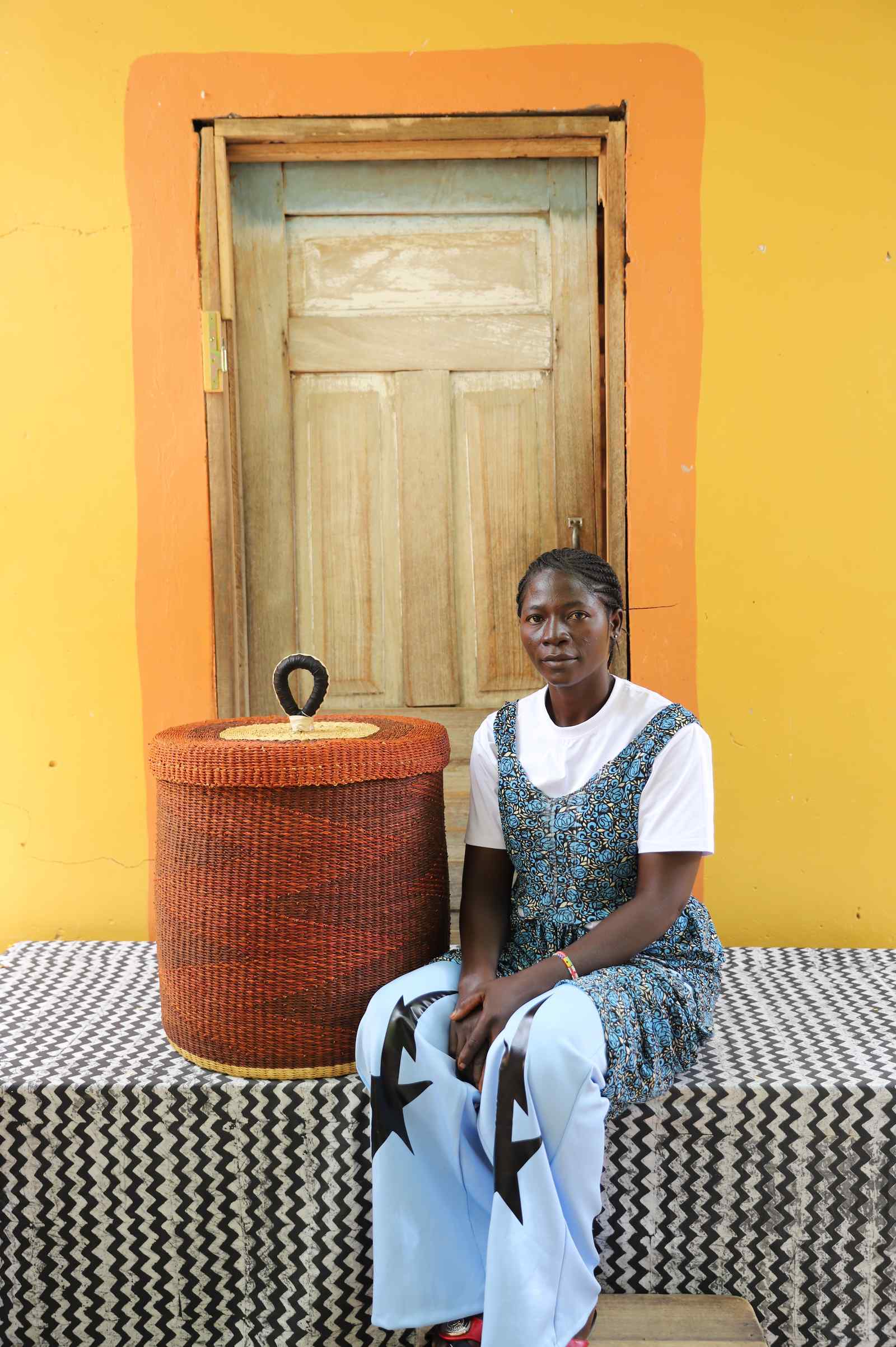 Handwoven Bolga laundry basket in dyed rust-red, beside a seated woman in blue and white.