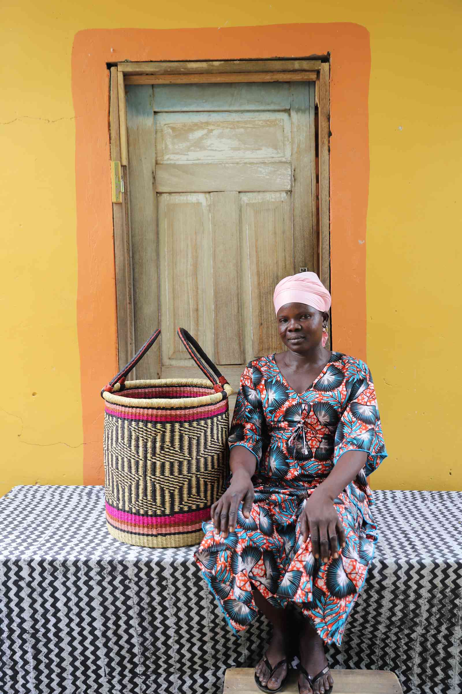 Small handwoven Bolga laundry basket in natural with red bands and dark weave, beside a seated woman.